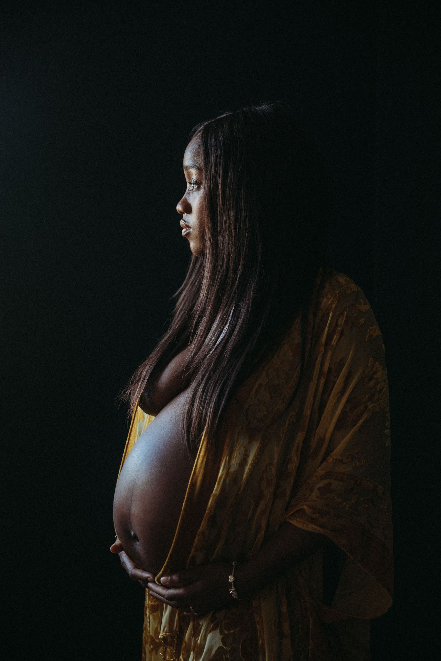 Black pregnant woman wearing yellow robe looking away from the camera by pregnancy photographer london sarah stirk