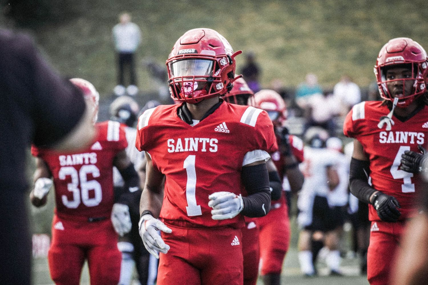 Saints football players in red uniforms warm up on the field, with player number 1 leading the team.