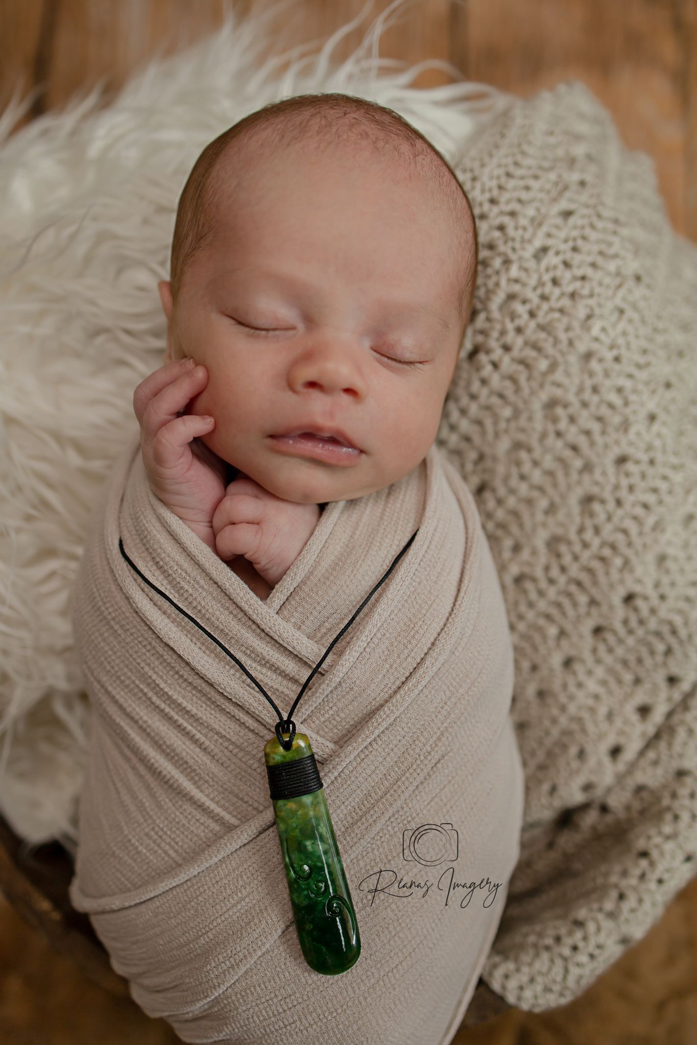 Sleeping newborn swaddled in cream wrap with green pendant necklace resting in fluffy white basket.