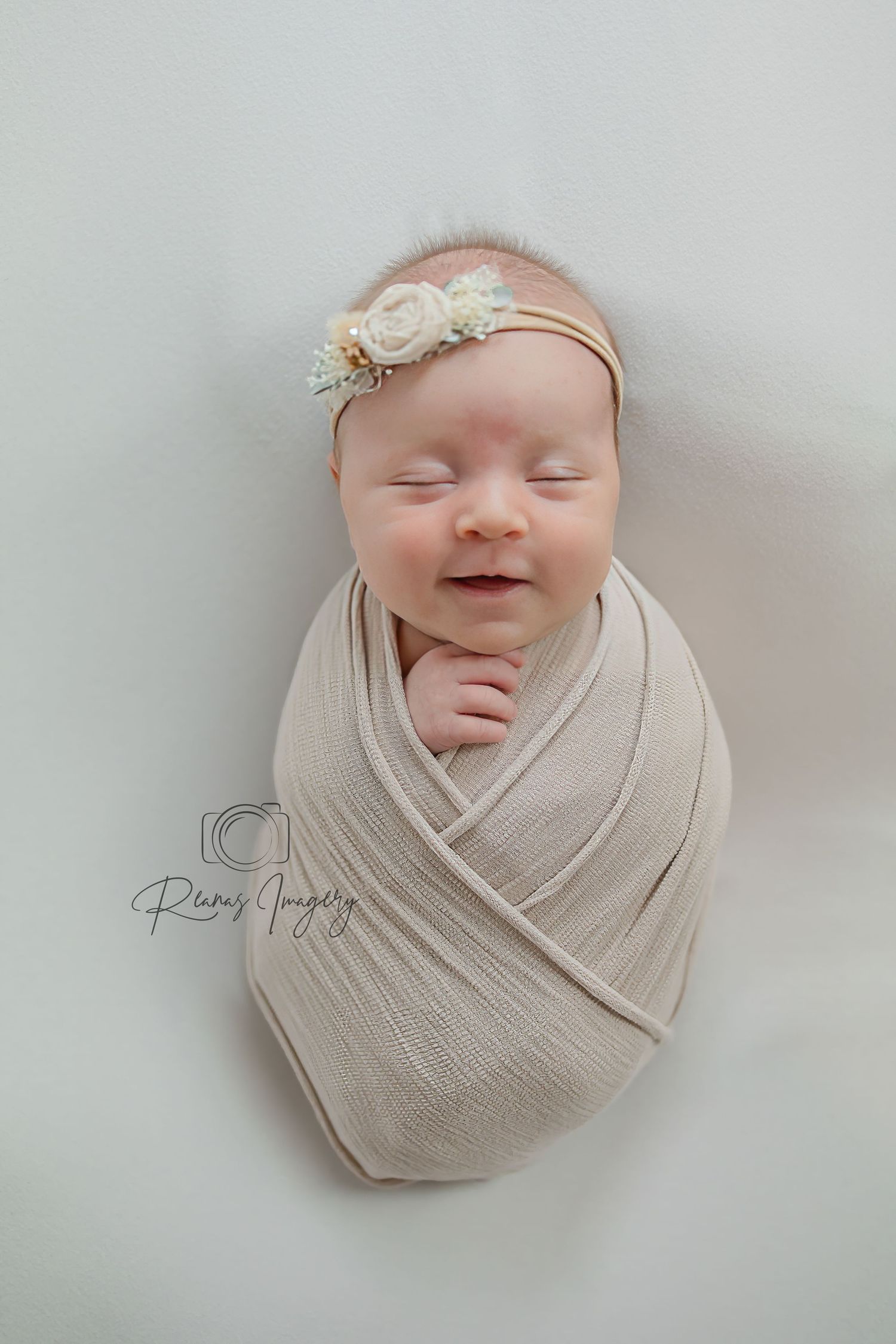 Smiling newborn baby swaddled in beige wrap wearing floral headband on white background.