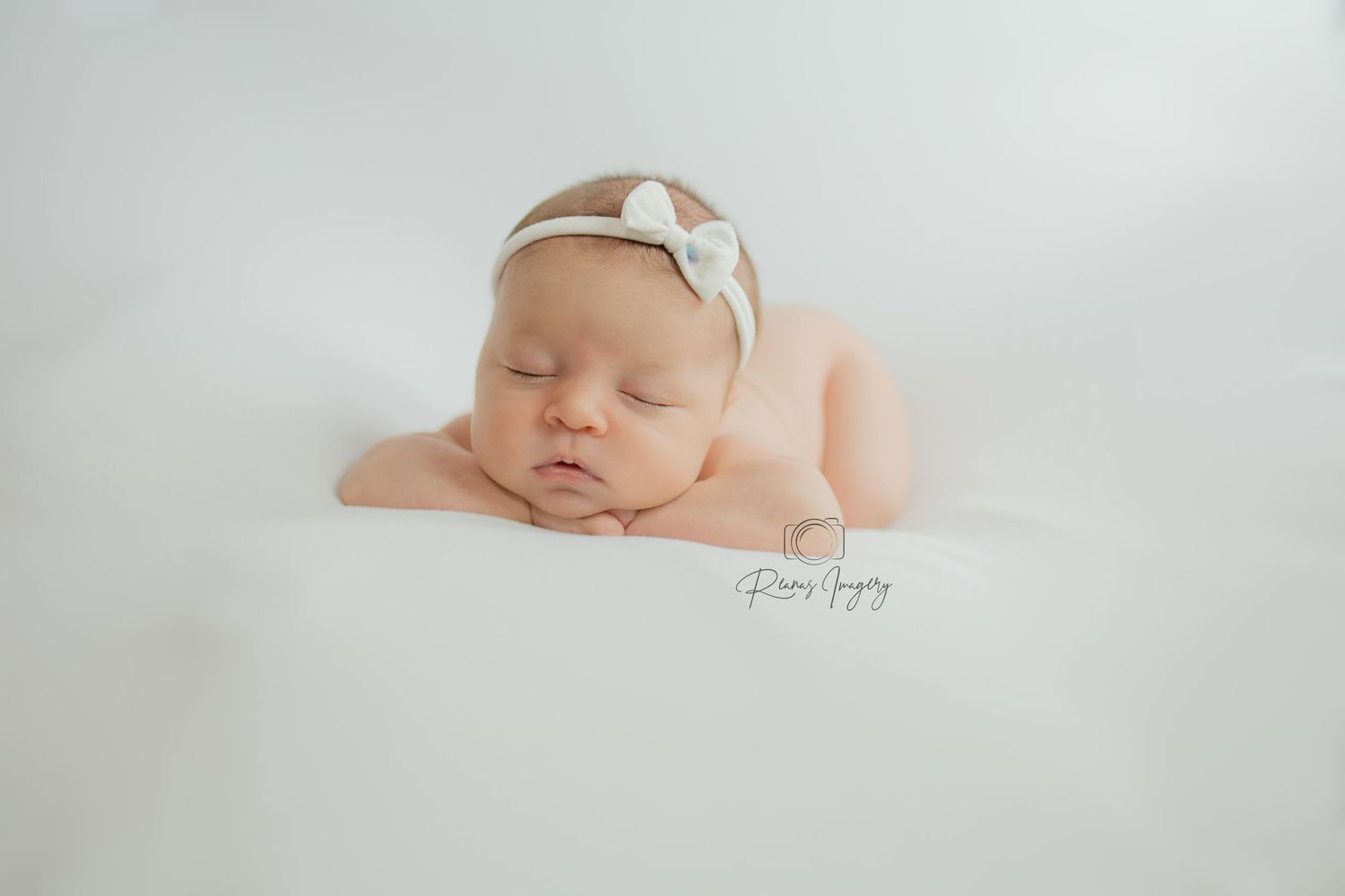 Peaceful newborn baby lying on white blanket wearing bow headband with hands under chin.