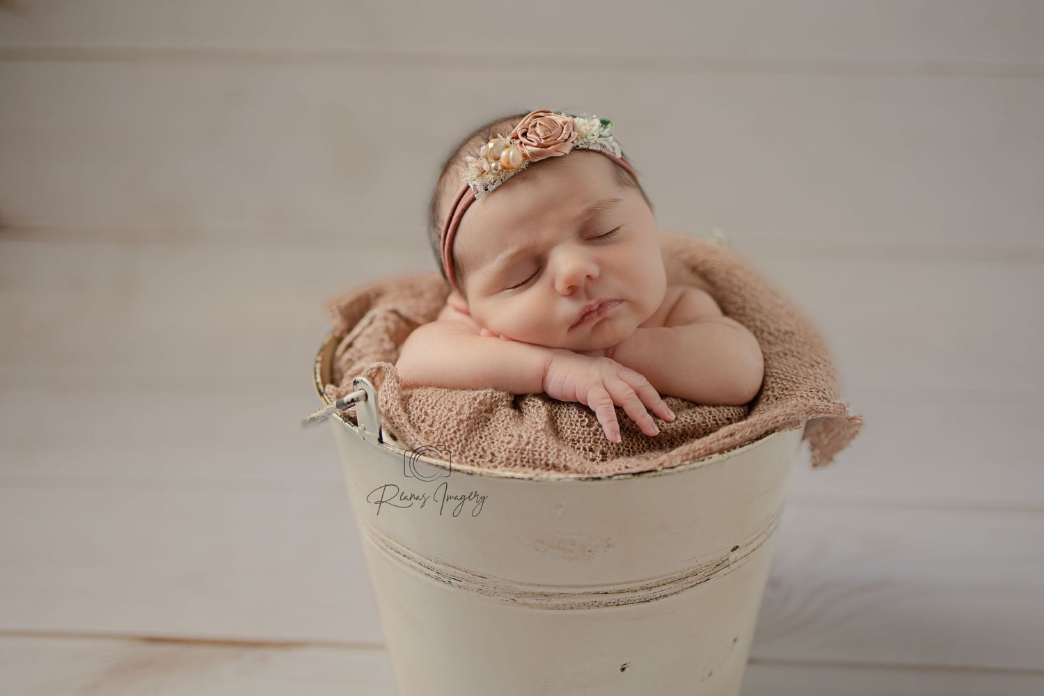Sleeping newborn in tan wrap resting in rustic white bucket with floral headband.