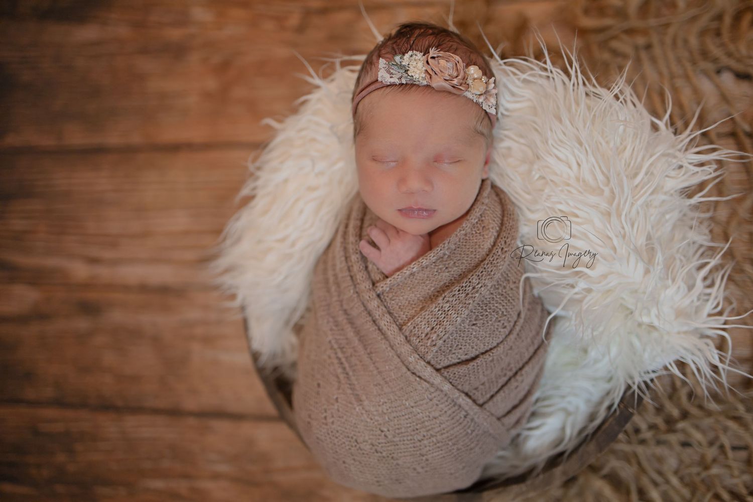 Newborn baby swaddled in brown knit wrap with floral headband in fluffy white basket.