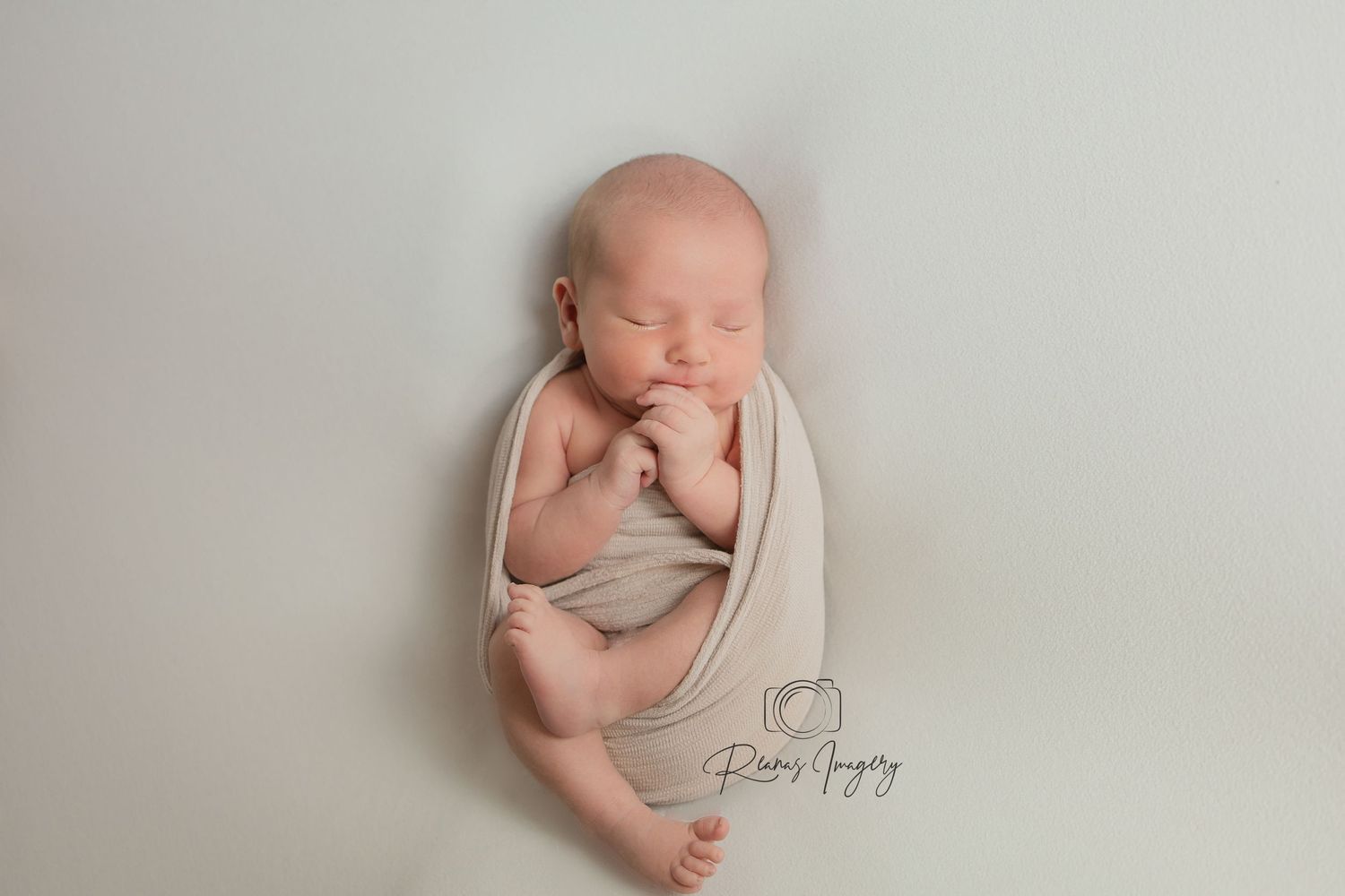 Sleeping newborn baby loosely swaddled in beige wrap lying on soft white background.