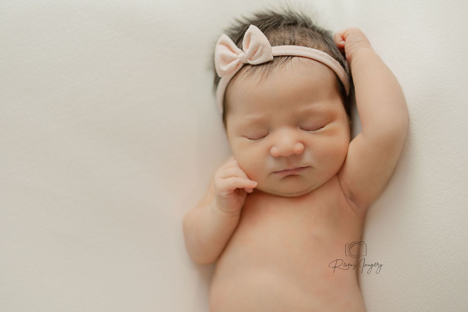 Peaceful newborn girl sleeping with arm raised wearing pink bow headband on white backdrop.