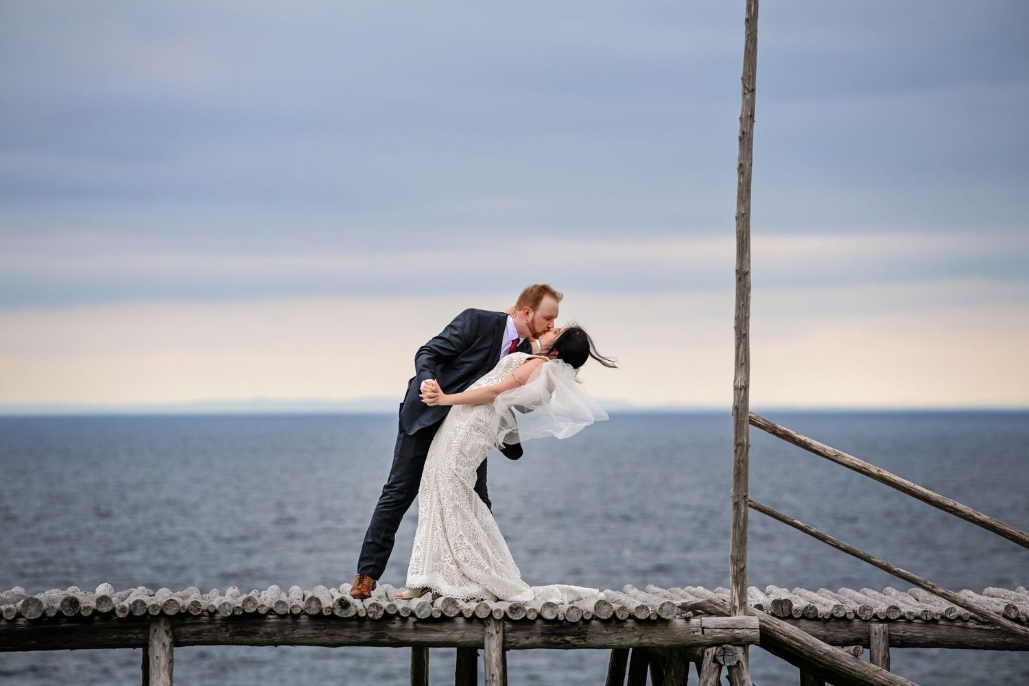 Romantic wedding couple in dramatic pose on pier overlooking ocean at sunset.
