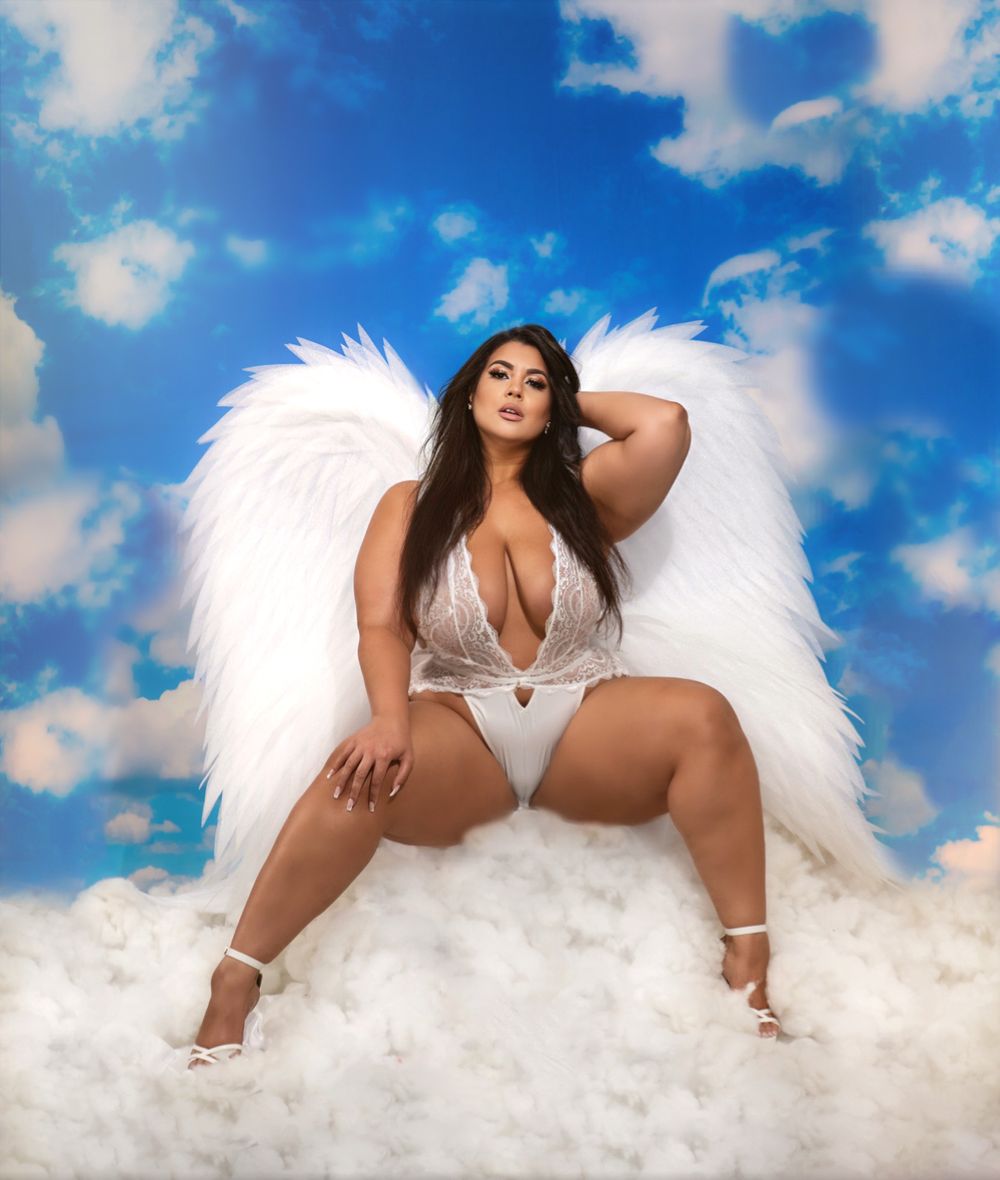 Model in white lingerie and angel wings poses on cloud backdrop against blue sky.