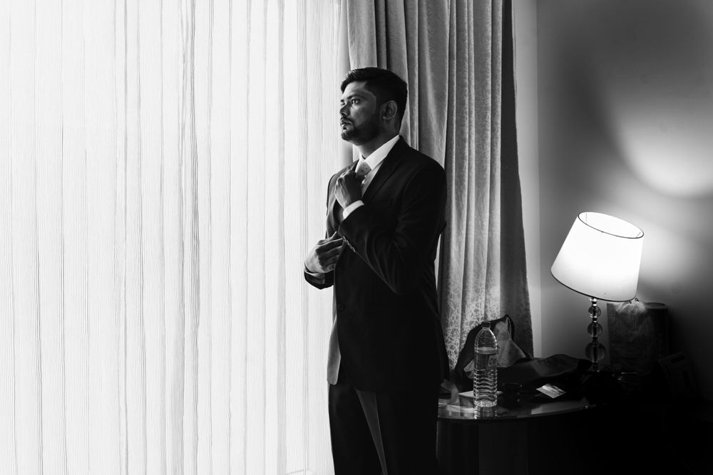 Black and white photo showing person adjusting tie near window with table lamp.