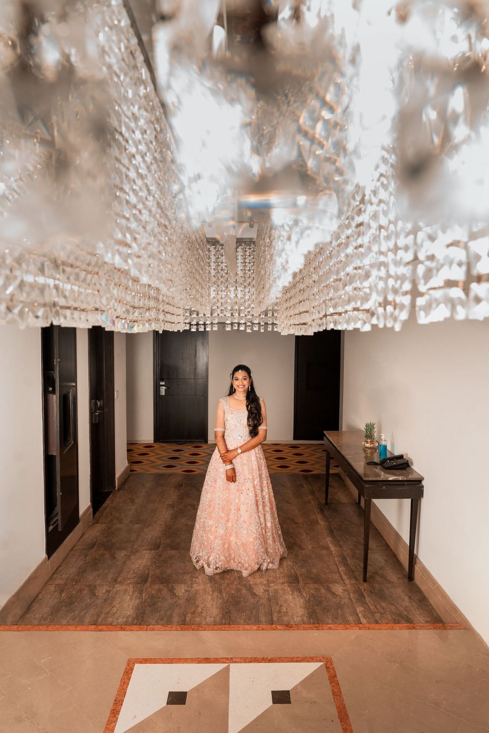 A figure in a pale pink lehenga stands beneath an ornate crystal ceiling in an elegant hallway.
