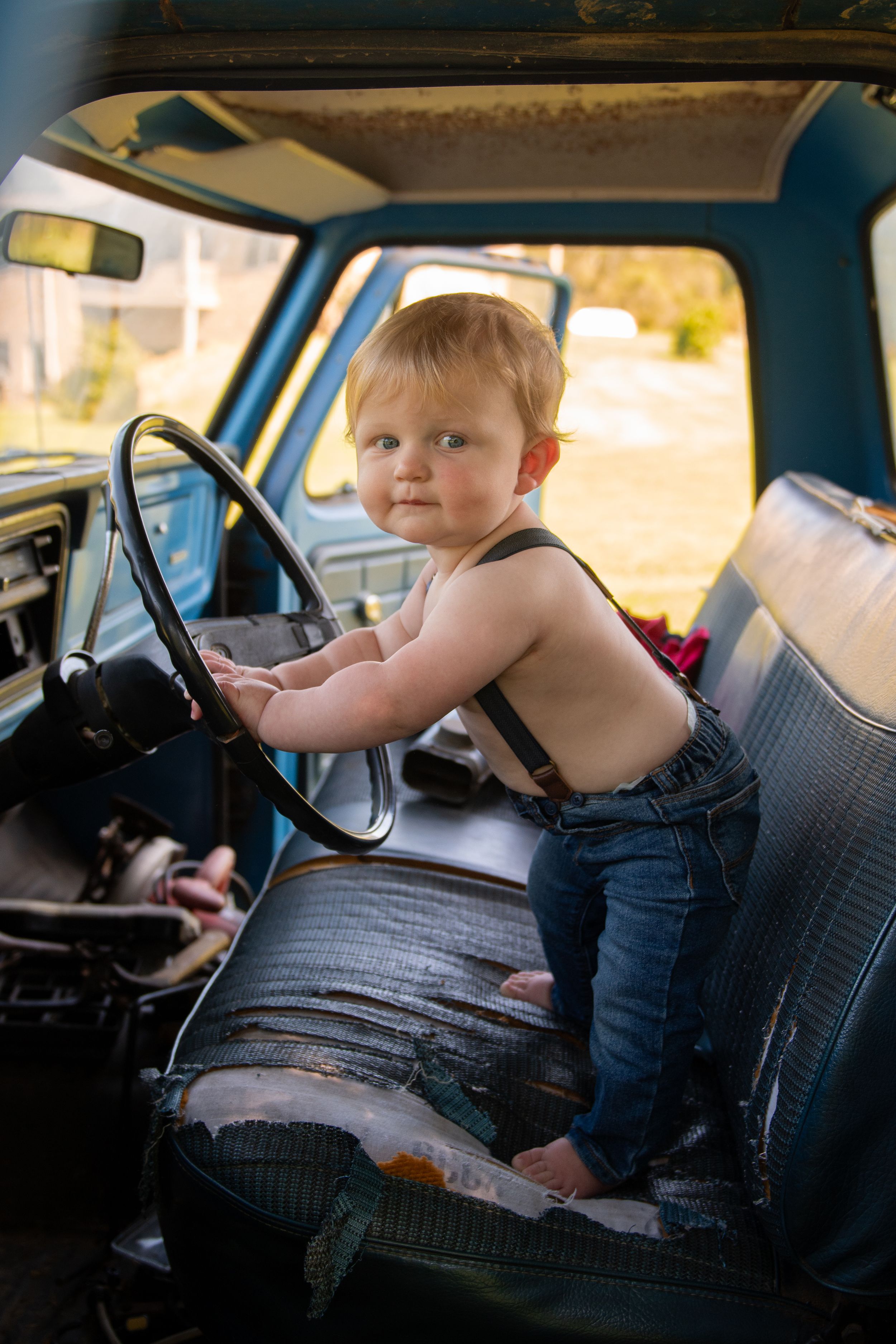 A small child sitting in the driver's seat of an older car, pretending to drive.