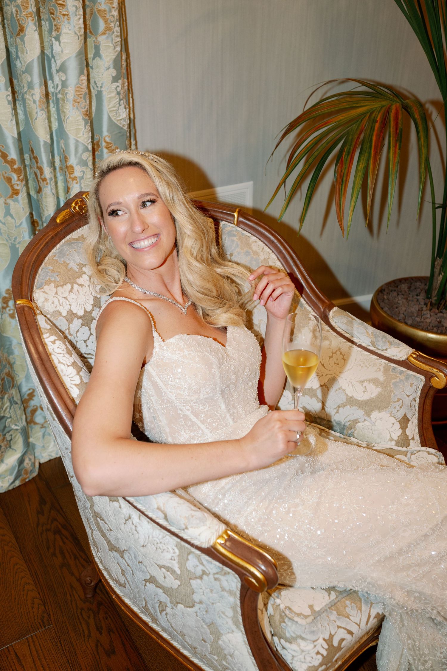 Bride in white lace dress relaxes on elegant vintage chair while holding champagne.