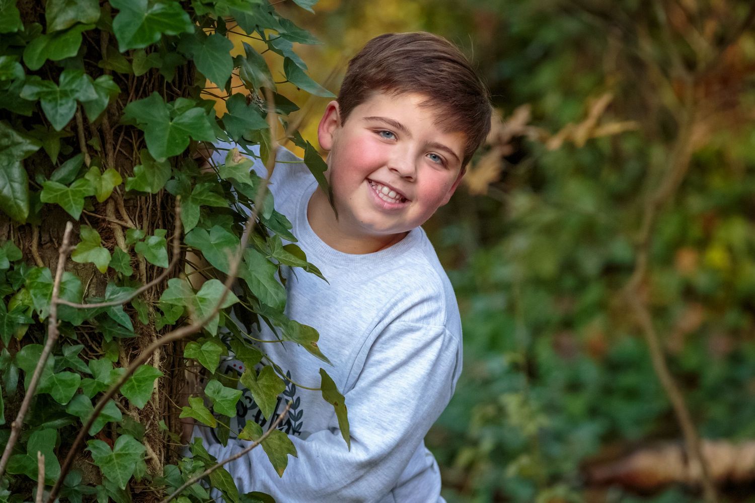 young boy leans around a large tree smiling during his mini photo session