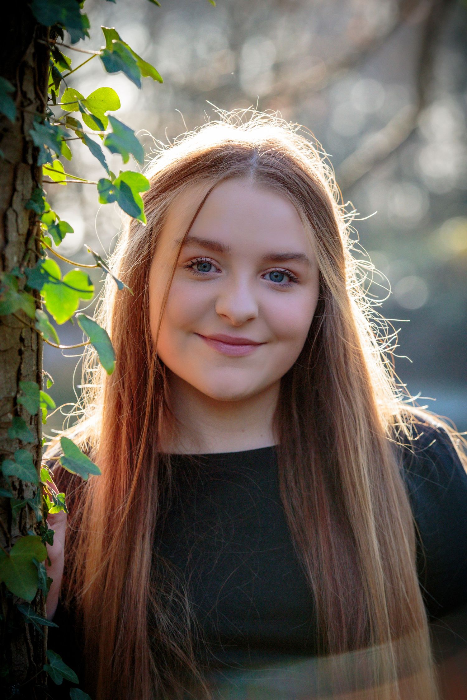 portrait of a teenage girl her hair backlit by the sun standing next to a tree covered in ivy