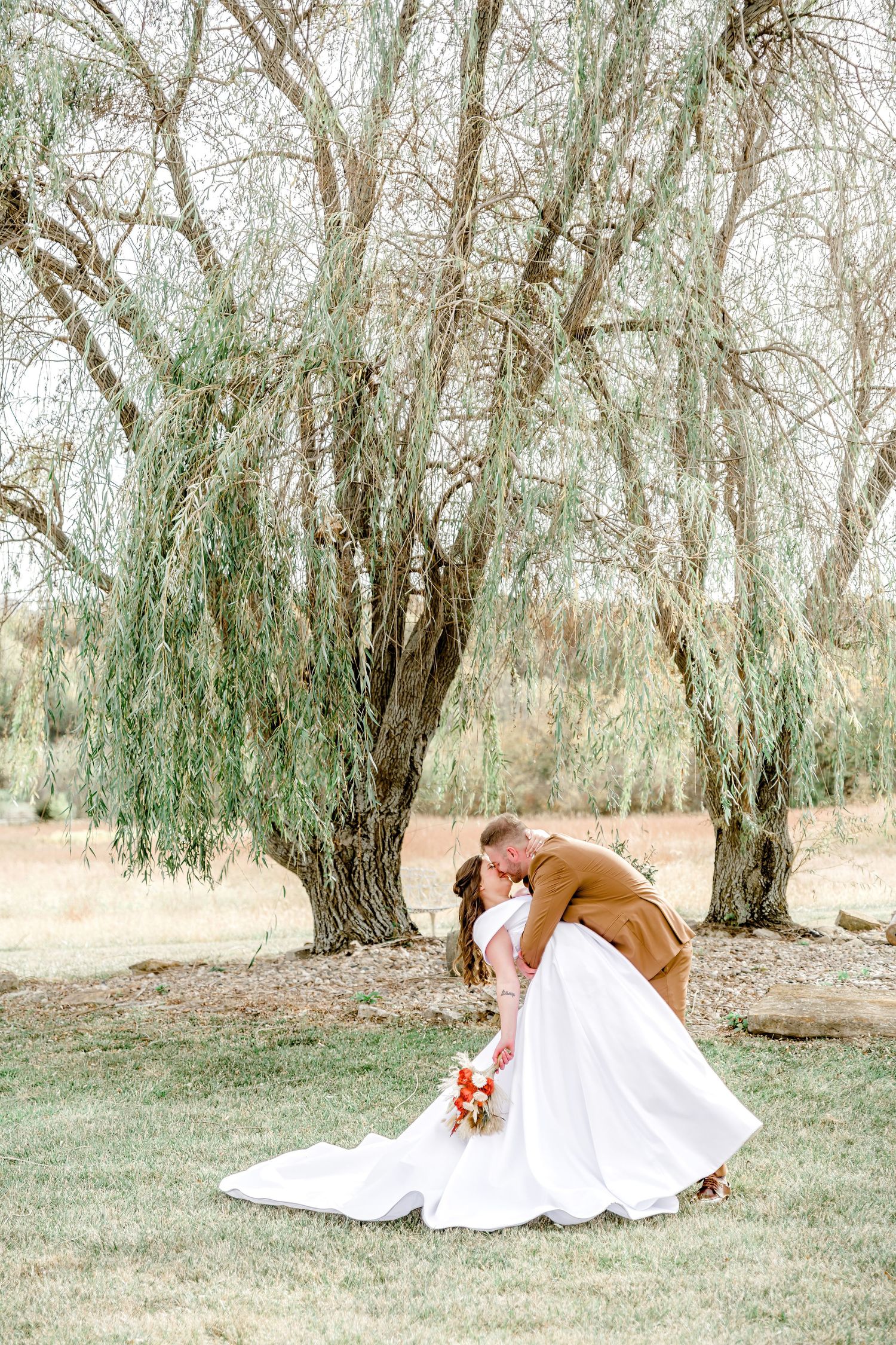 A romantic embrace under a weeping willow tree on a sunny summer day with a flowing white dress and bouquet by Stephanie Coulter Photography