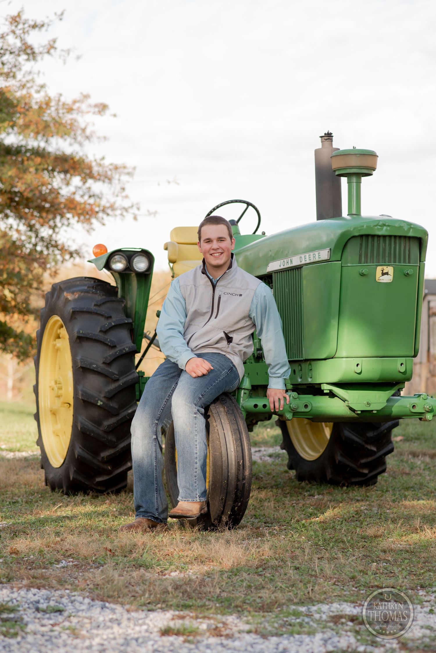 Person sits on large wheel of vintage John Deere tractor near stone building.