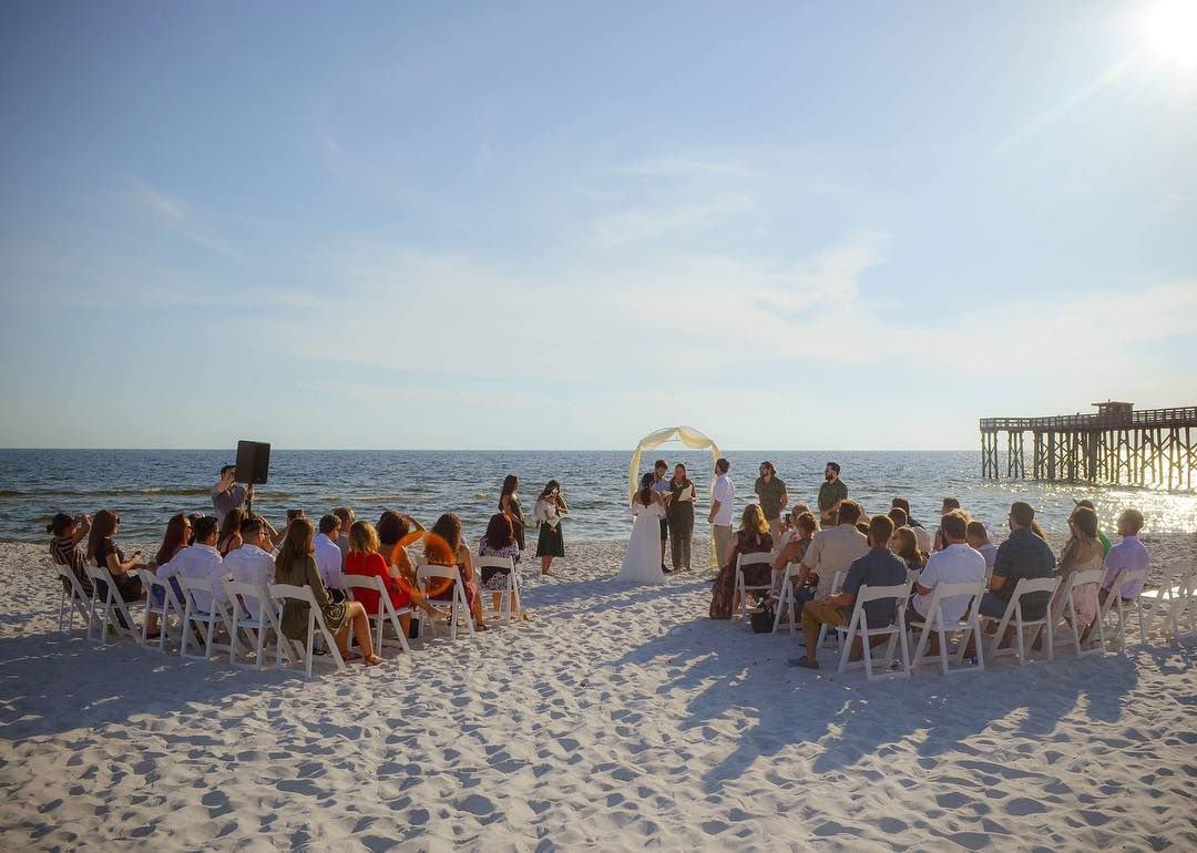 Outdoor beach wedding ceremony with guests seated in white chairs on white sand near a pier at sunset.