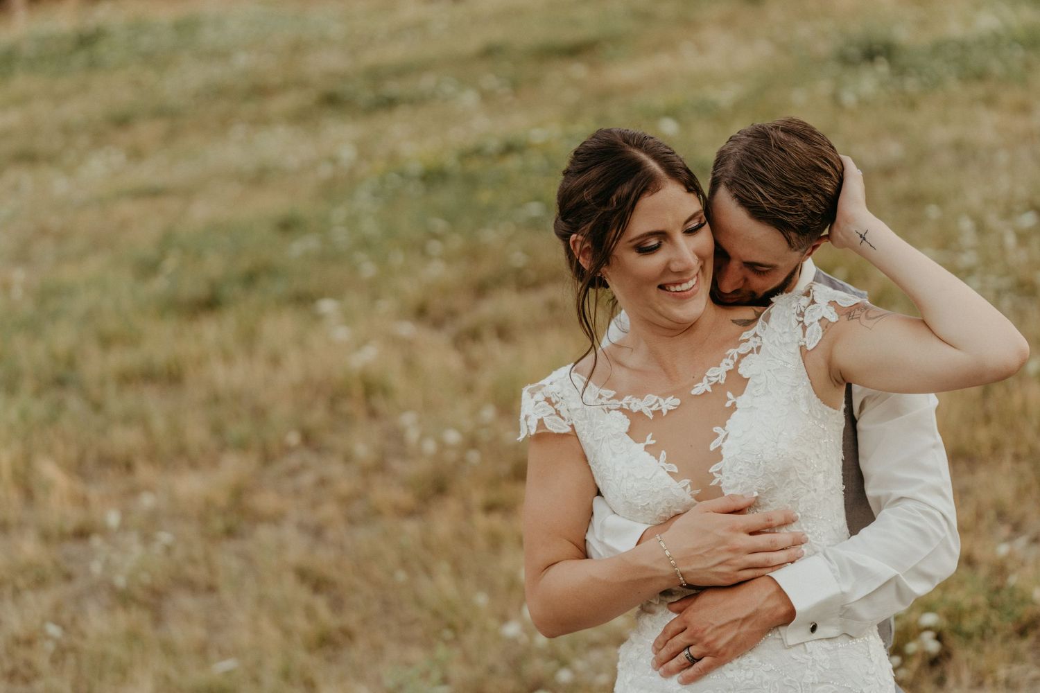 Romantic couple embracing in lace wedding attire against rustic outdoor backdrop.