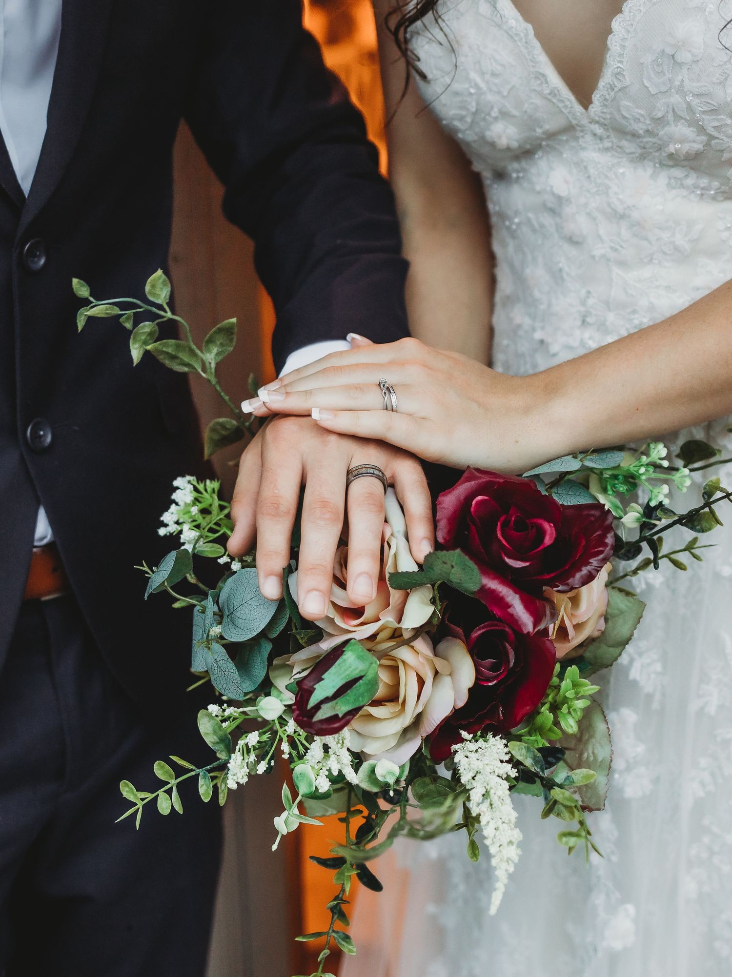 Close-up of wedding rings and hands with deep red roses and greenery bouquet.