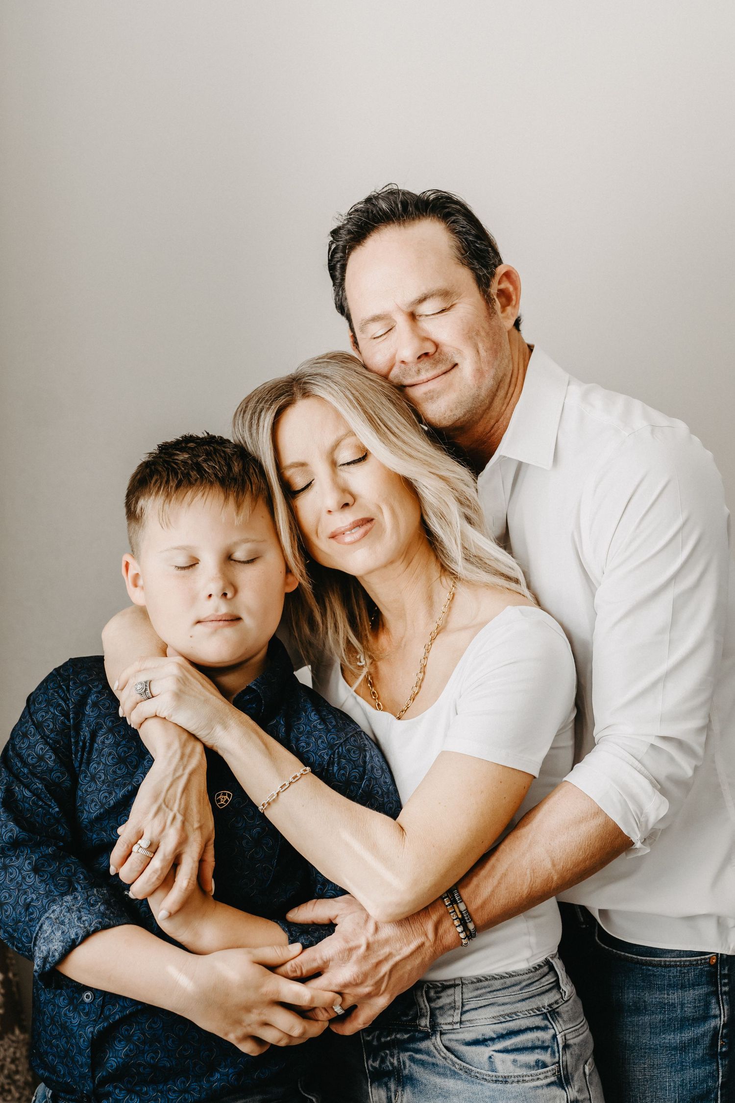 Family portrait of parents and young son embracing with eyes closed against a light grey background.
