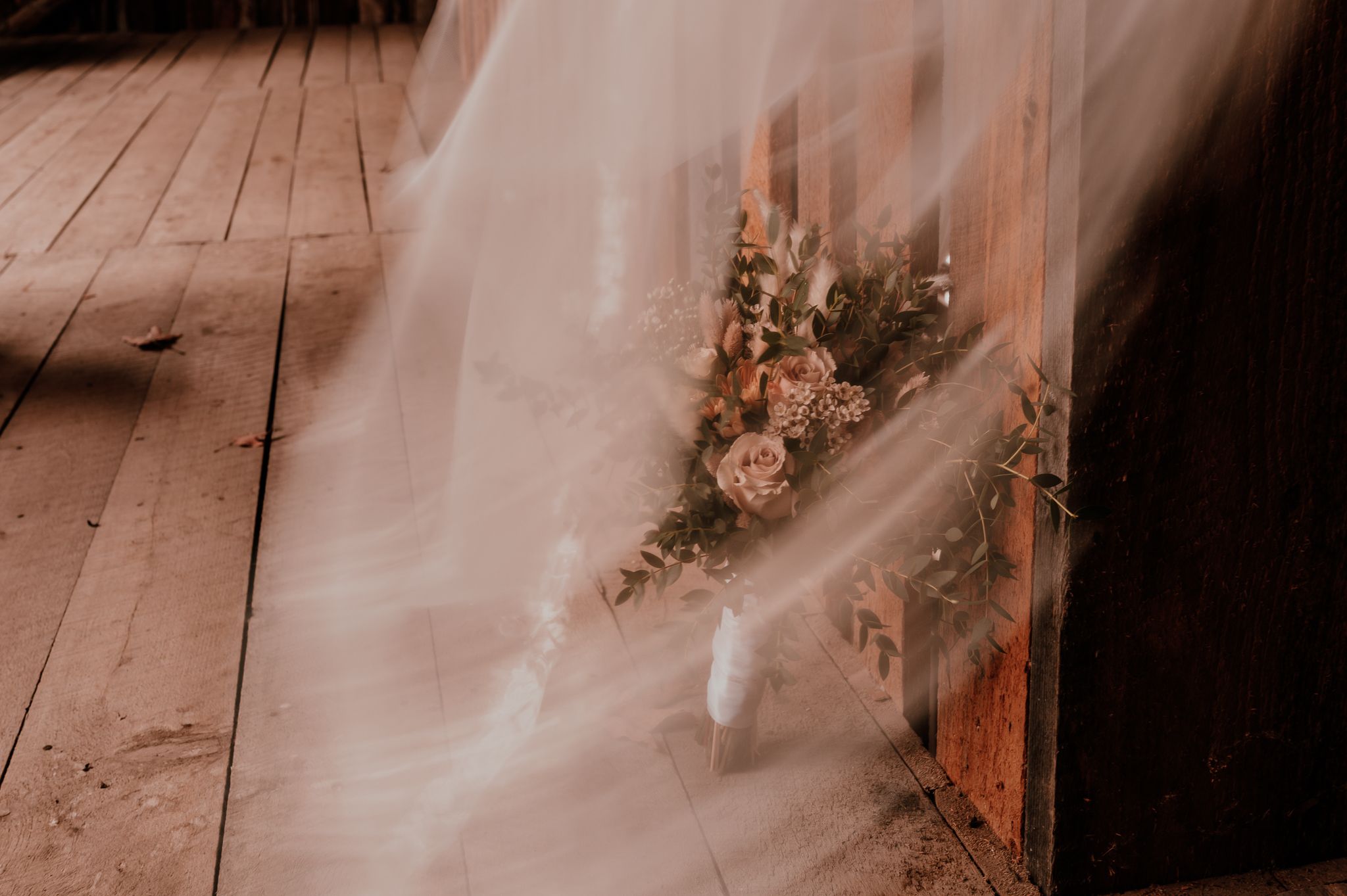 A delicate veil drapes over dried floral arrangements on a rustic wooden floor in moody lighting.