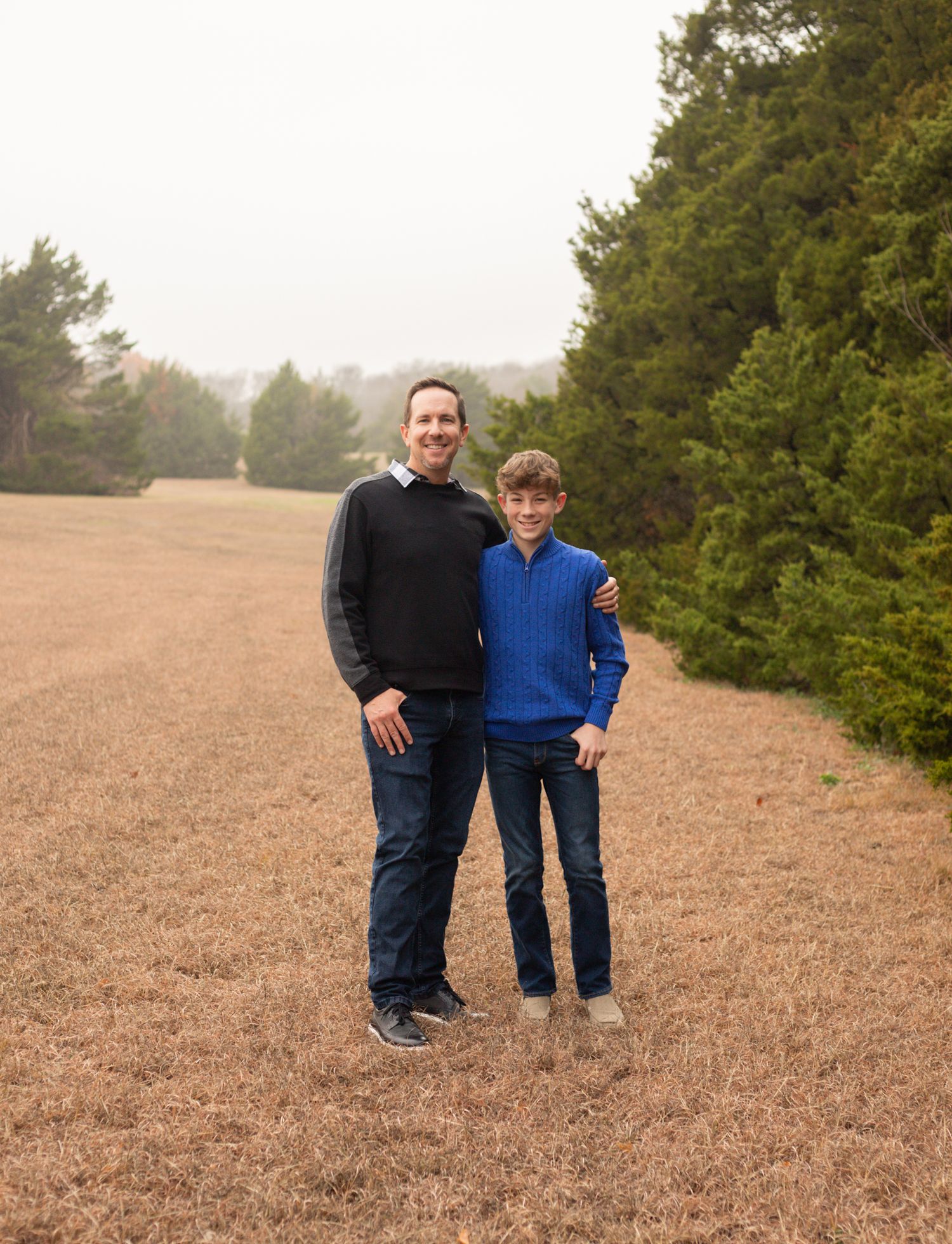 Father and son standing together smiling in an open field with evergreen trees in the background.