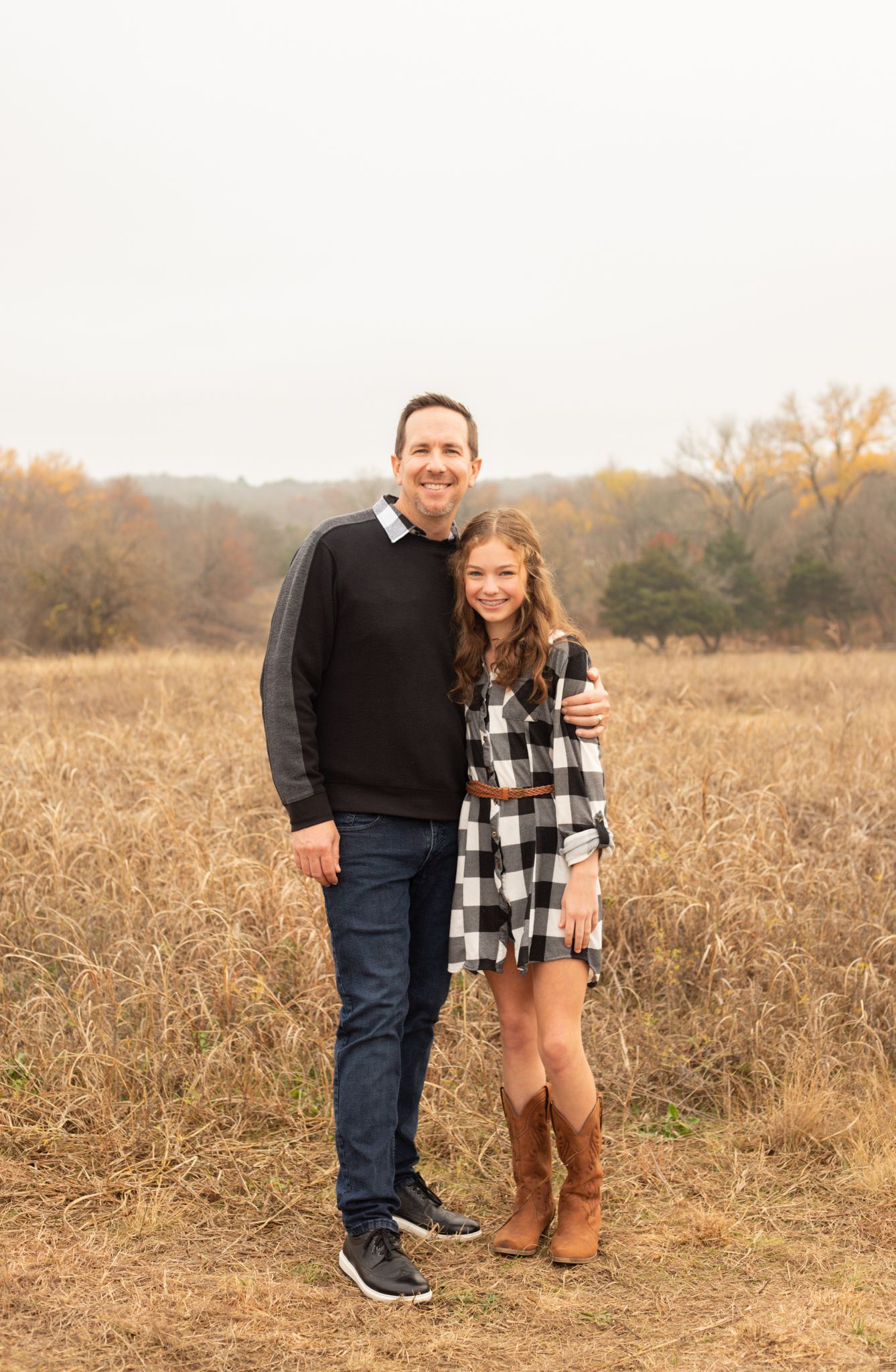 Father and daughter in plaid dress and cowboy boots posing in autumn field.