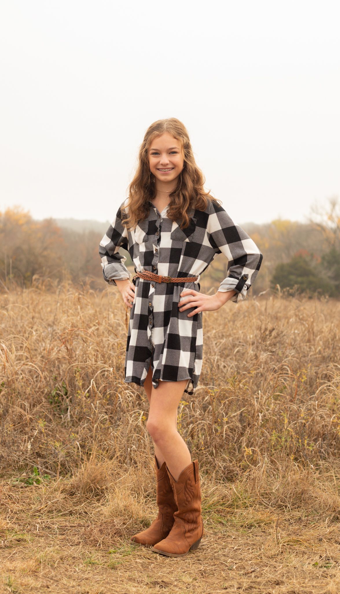 Smiling girl in plaid romper and cowboy boots poses in golden autumn field.