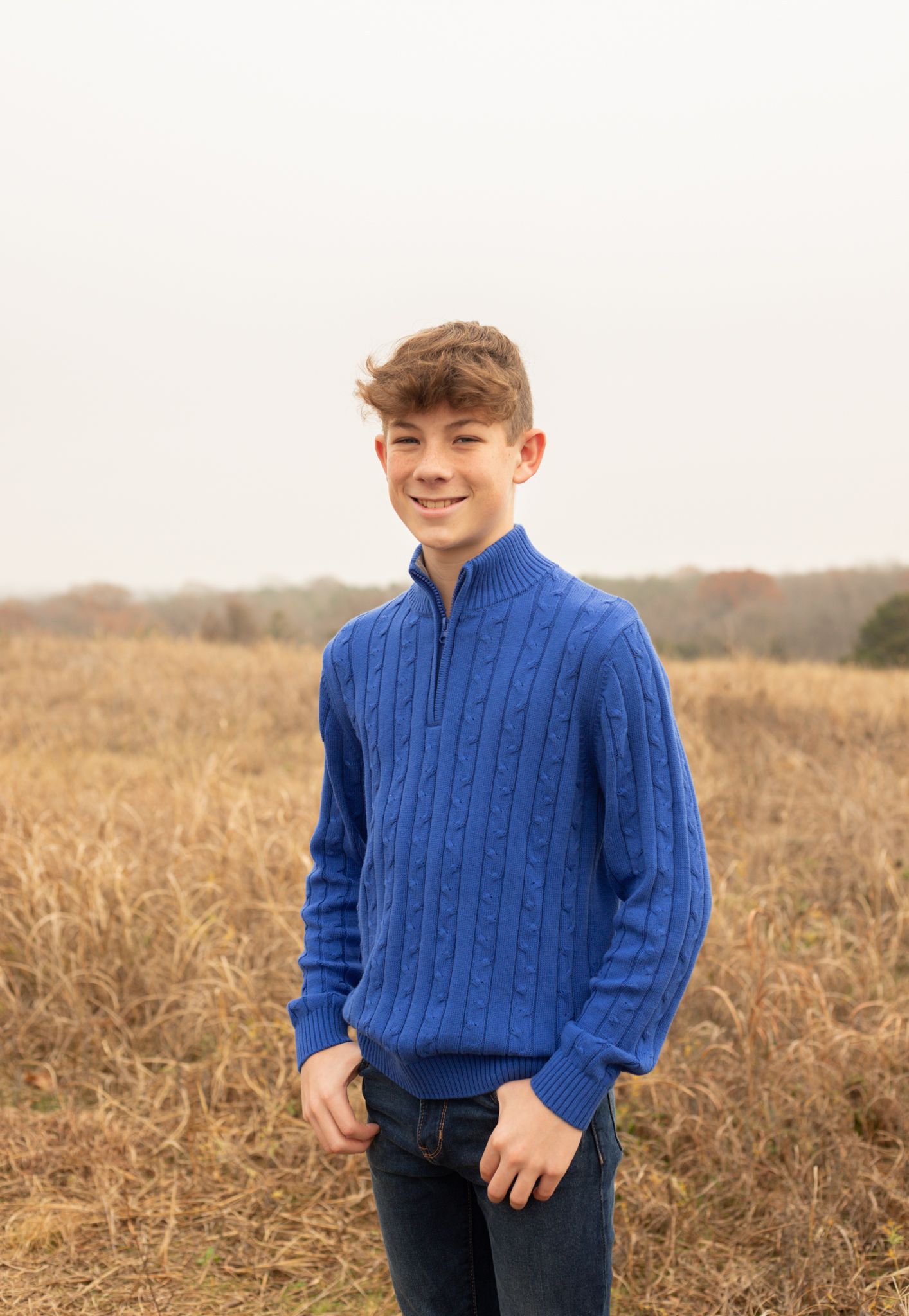 Teenage boy in blue cable-knit sweater smiling in dry autumn grass field.