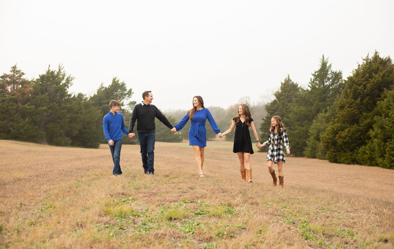 A happy family of five holds hands while walking together across an open grassy field outdoors.