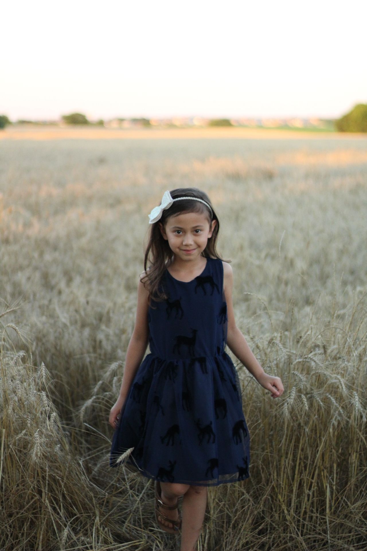 Young girl in navy blue dress with bow headband standing in golden wheat field at sunset.