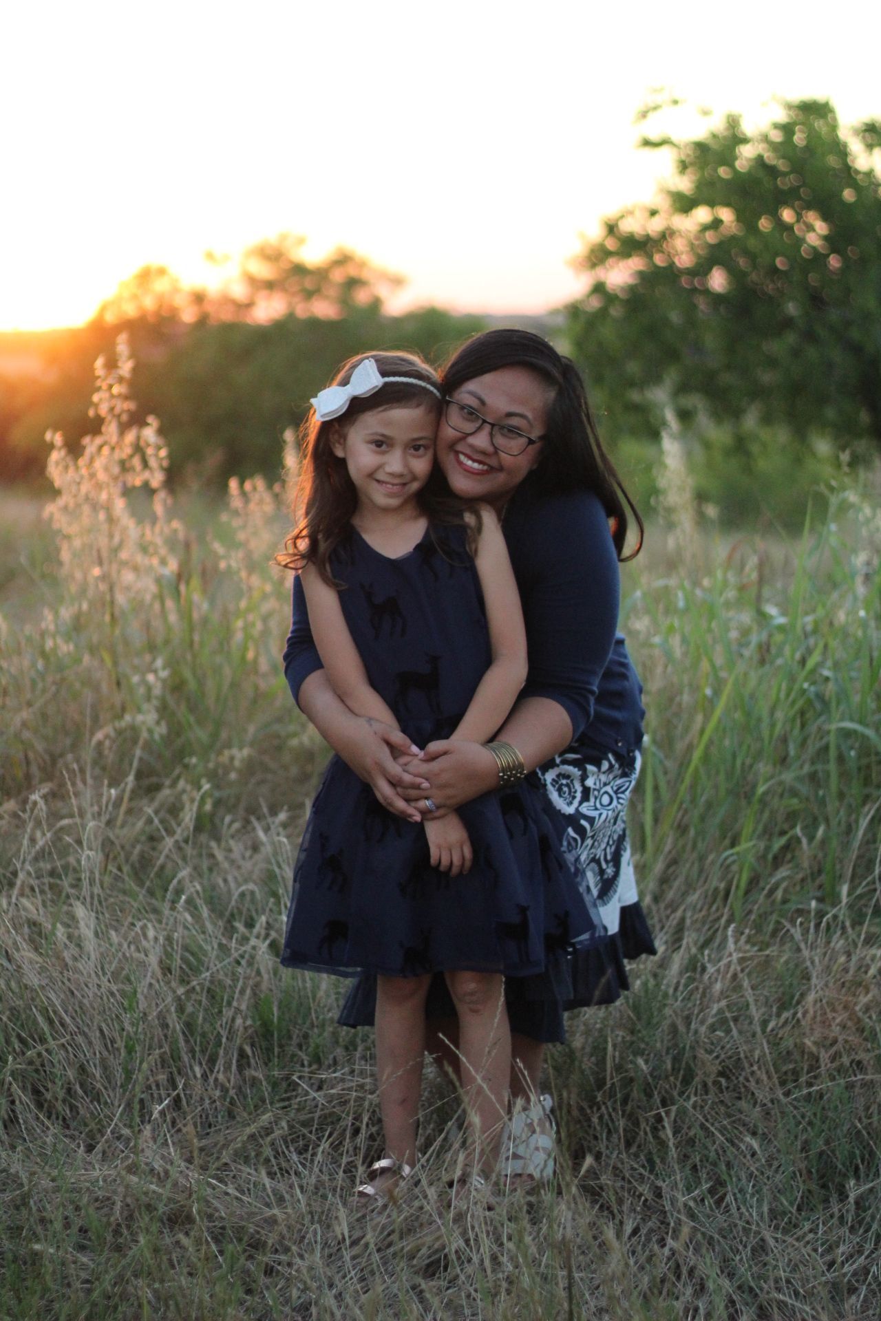 Mother and young daughter in matching navy dresses embracing in a golden sunset field.