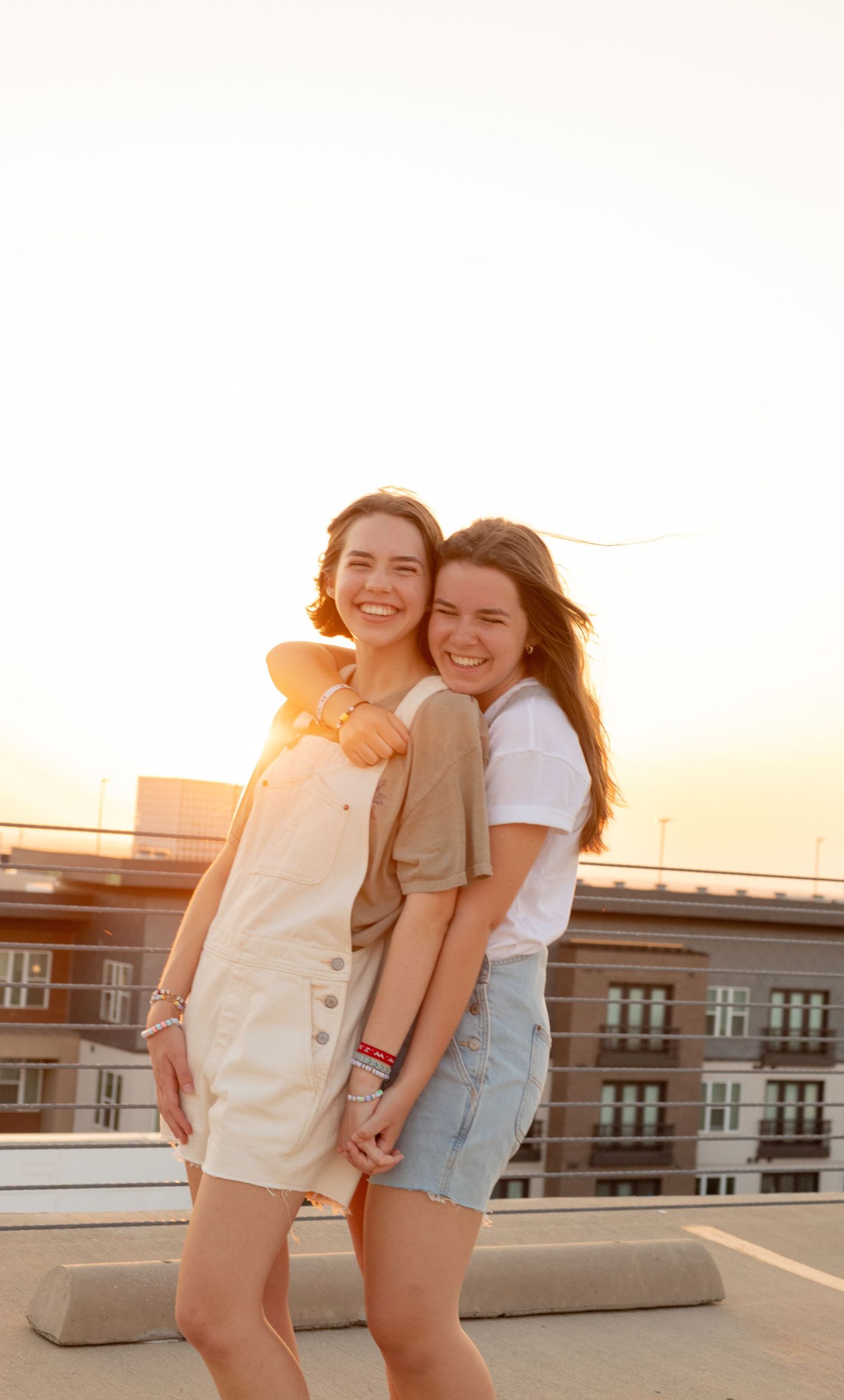 Two smiling young women embracing on a rooftop during a beautiful golden hour sunset.