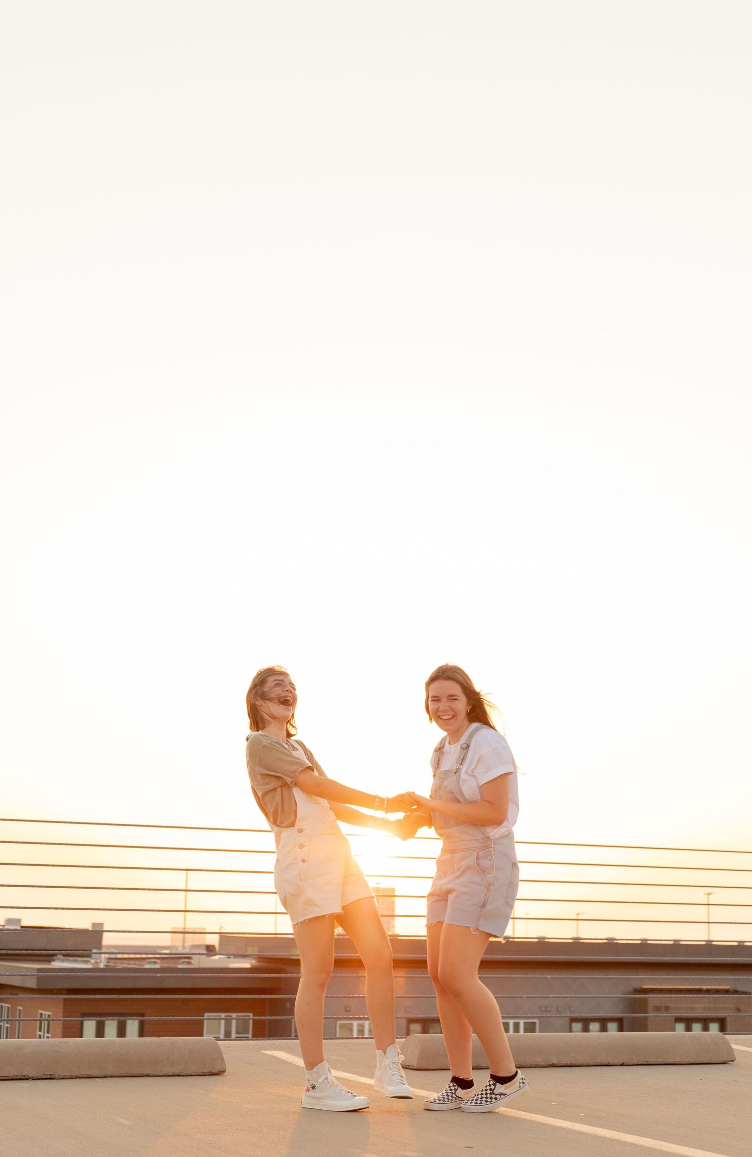 Two young women holding hands and smiling on a rooftop during a vibrant golden sunset.