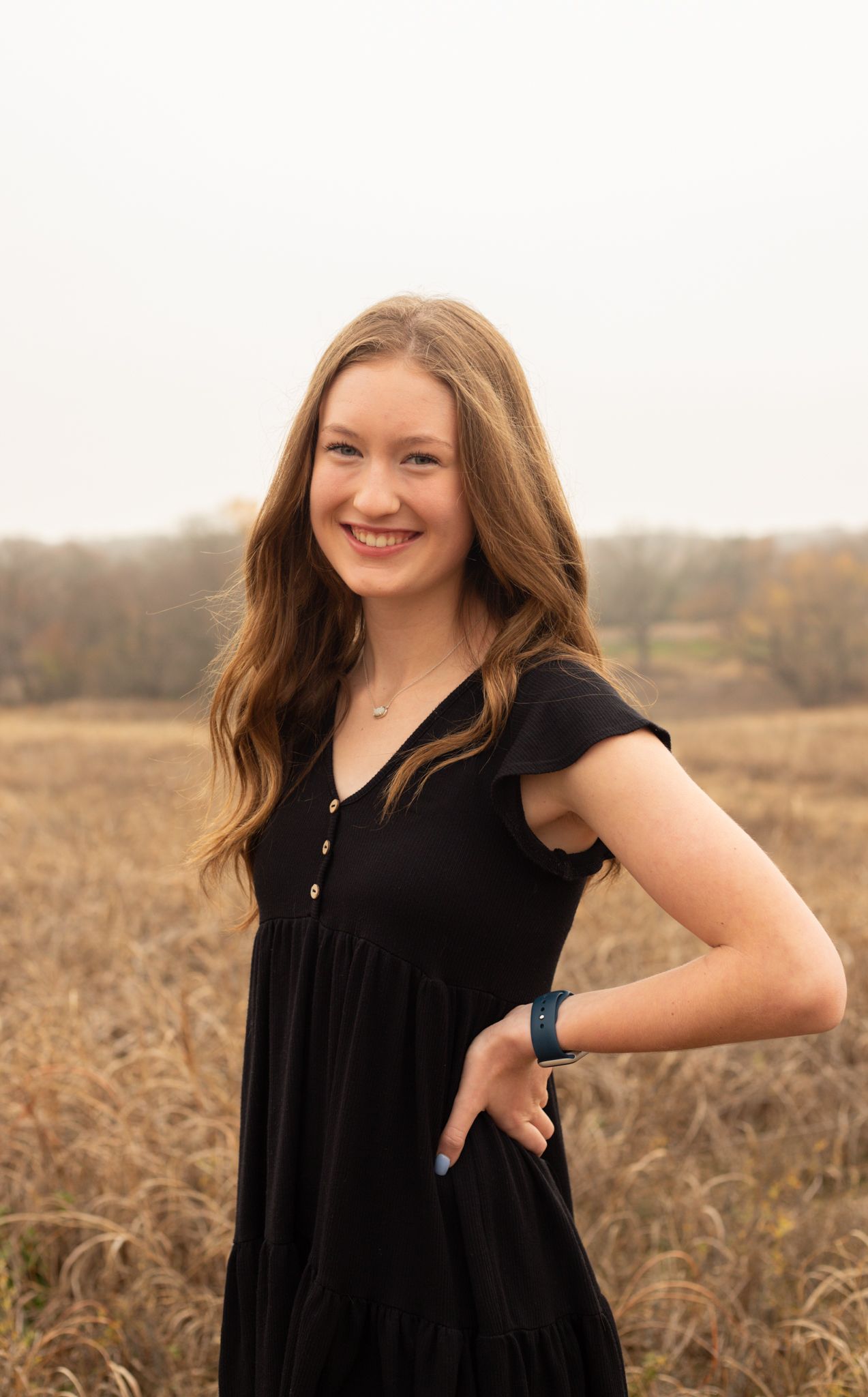 Young woman in black dress poses smiling in autumn field with golden grass backdrop.