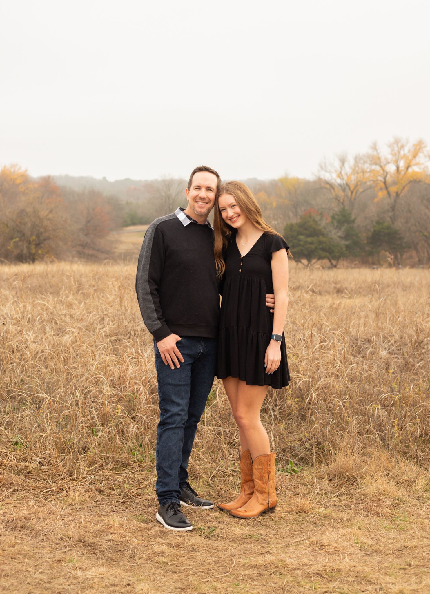 Father and teenage daughter in black dress smiling together in fall outdoor setting.