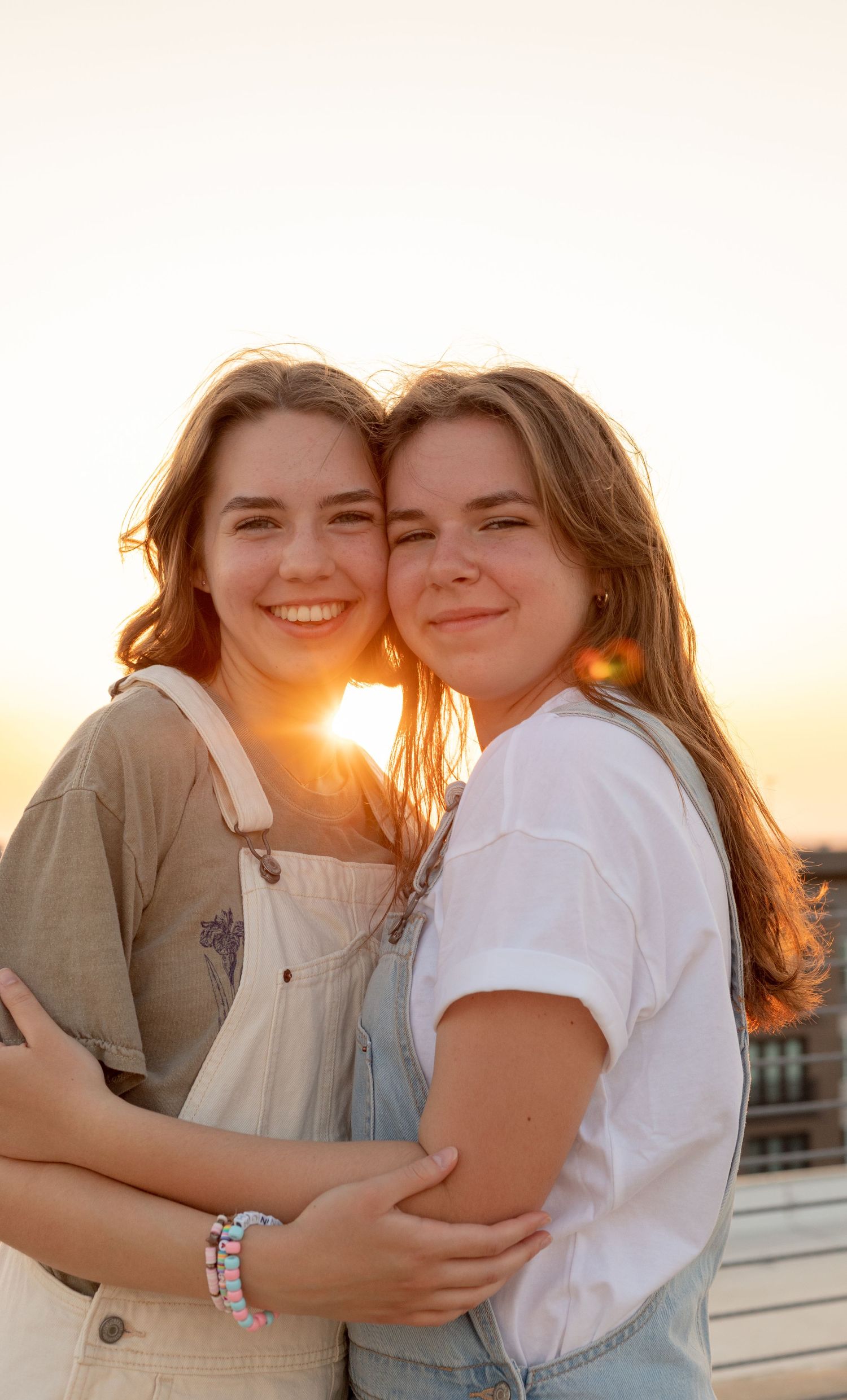 Two smiling teenage girls hugging on a rooftop at golden hour sunset.