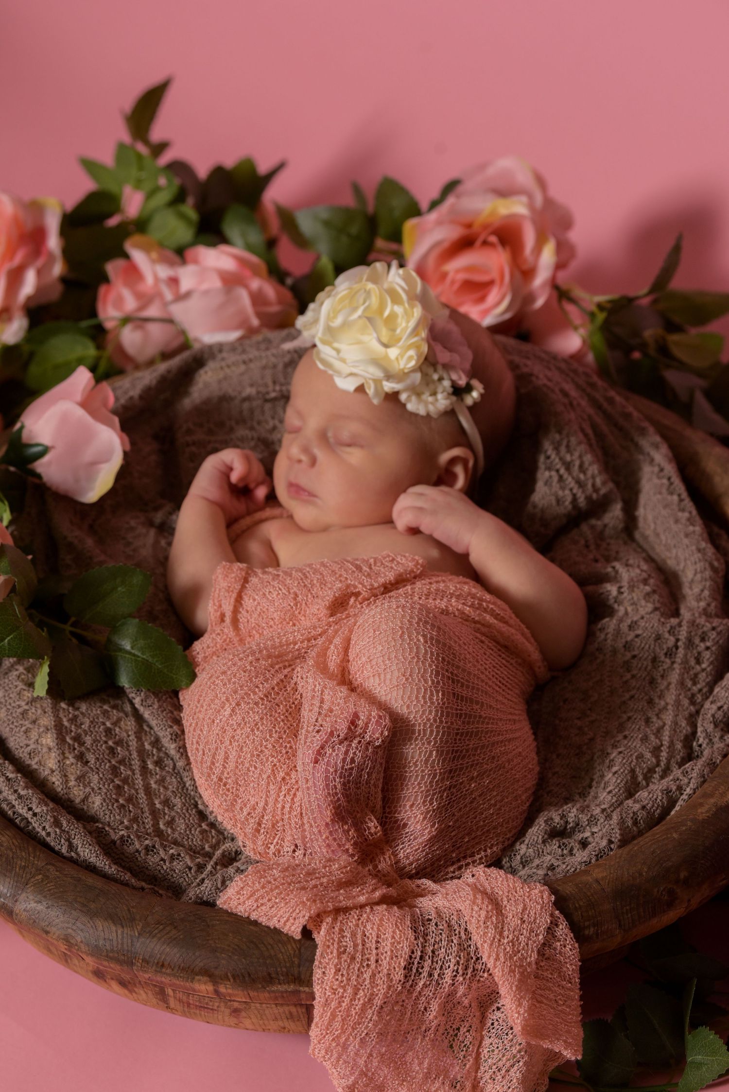 A newborn baby girl sleeps in a wooden bowl surrounded by pink roses and soft wraps.
