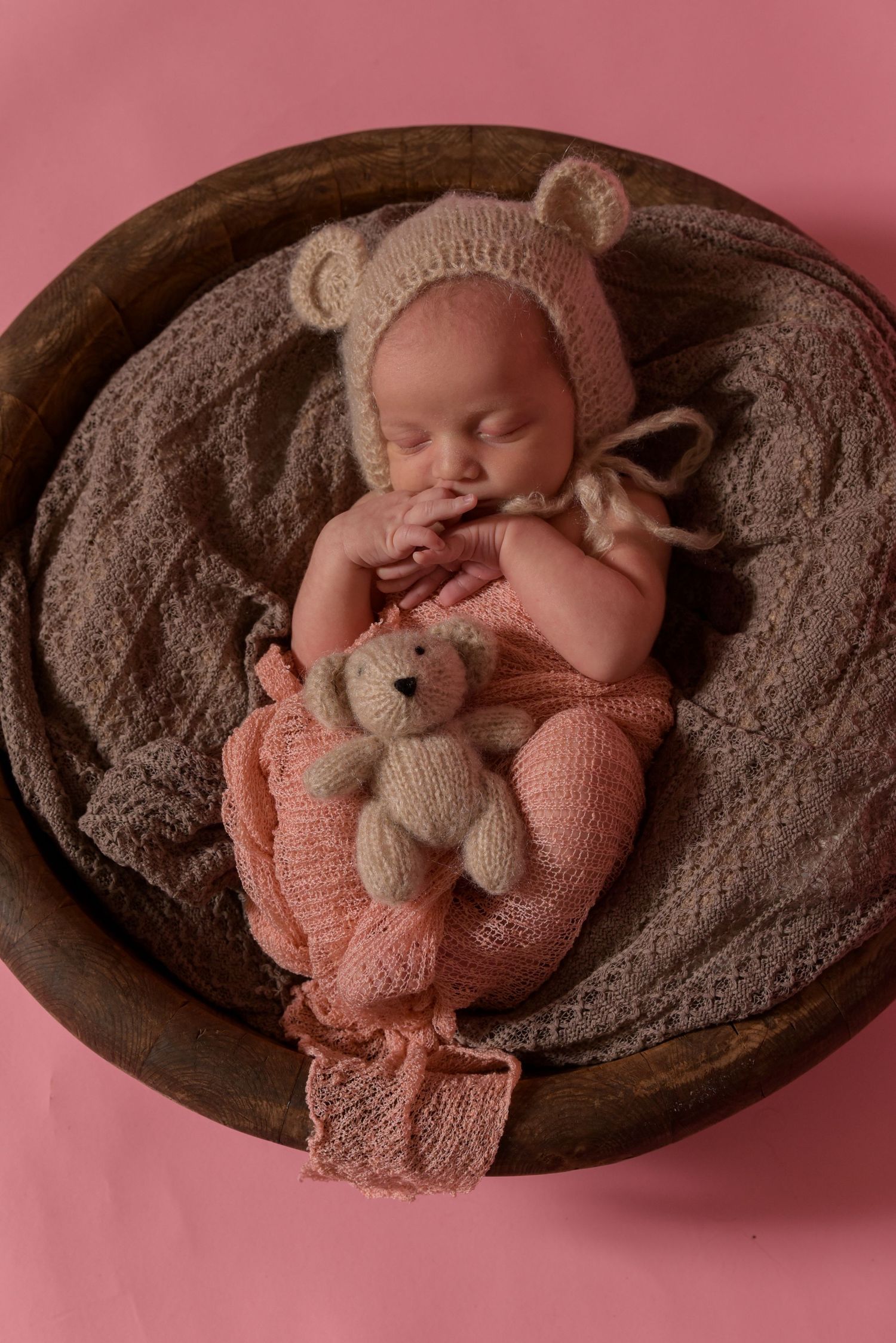 A sleeping newborn in a bear bonnet and pink knit outfit holds a tiny teddy bear.