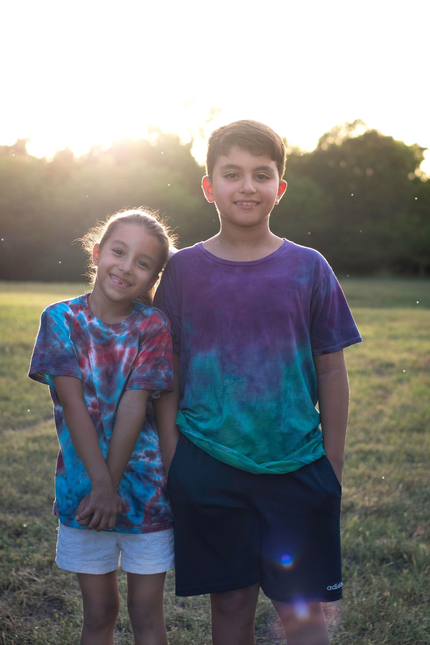 Two siblings in colorful tie-dye shirts smiling together in a sunlit green field.