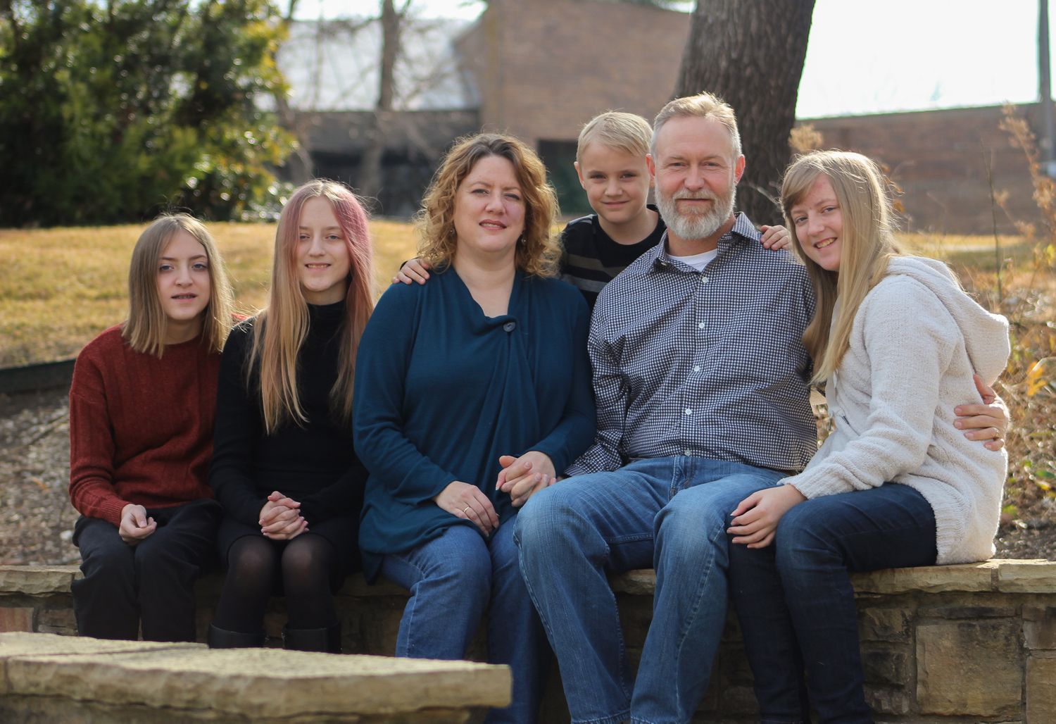 Family of six smiling together while seated on a stone wall in a park setting.