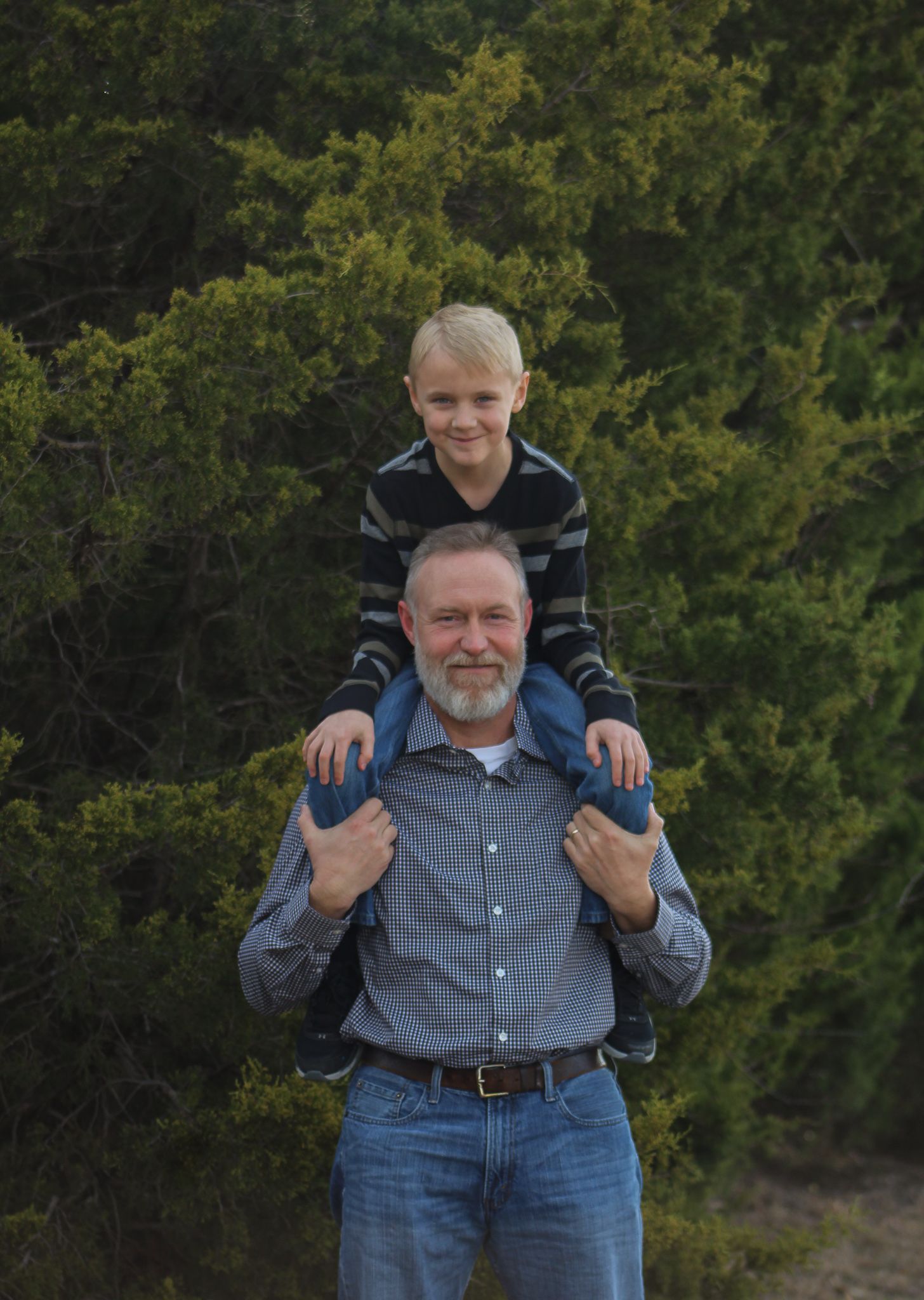 Smiling grandfather carrying young blonde grandson on his shoulders outdoors near green trees.
