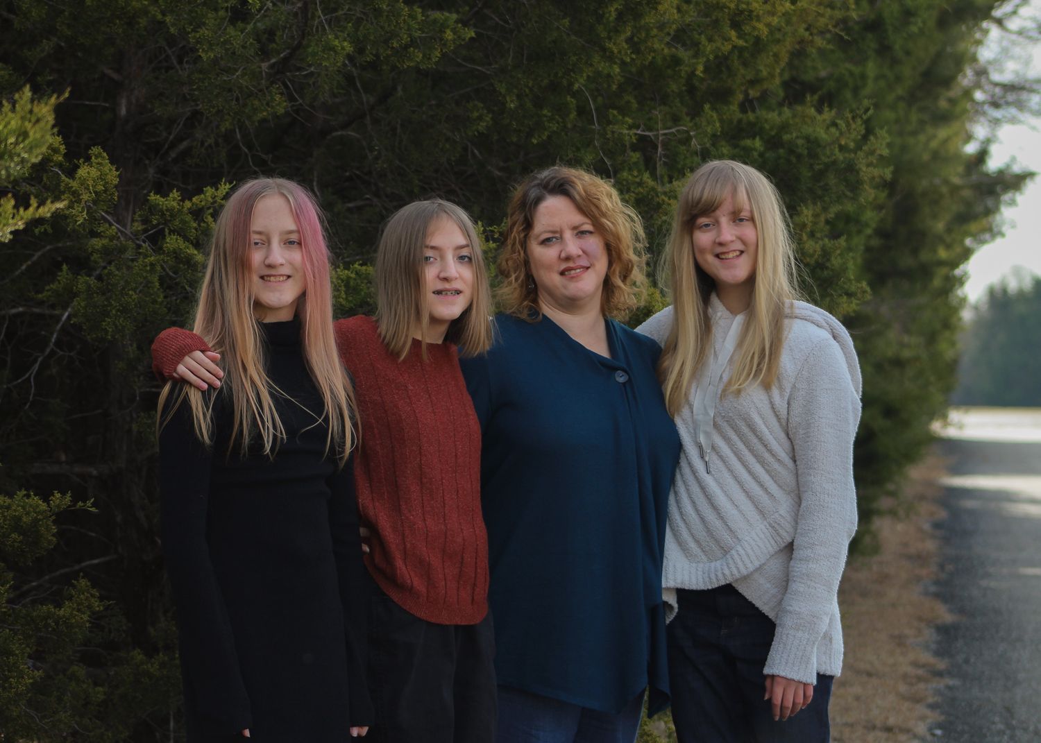 Mother and three teenage daughters posing together along a tree-lined road outdoors.