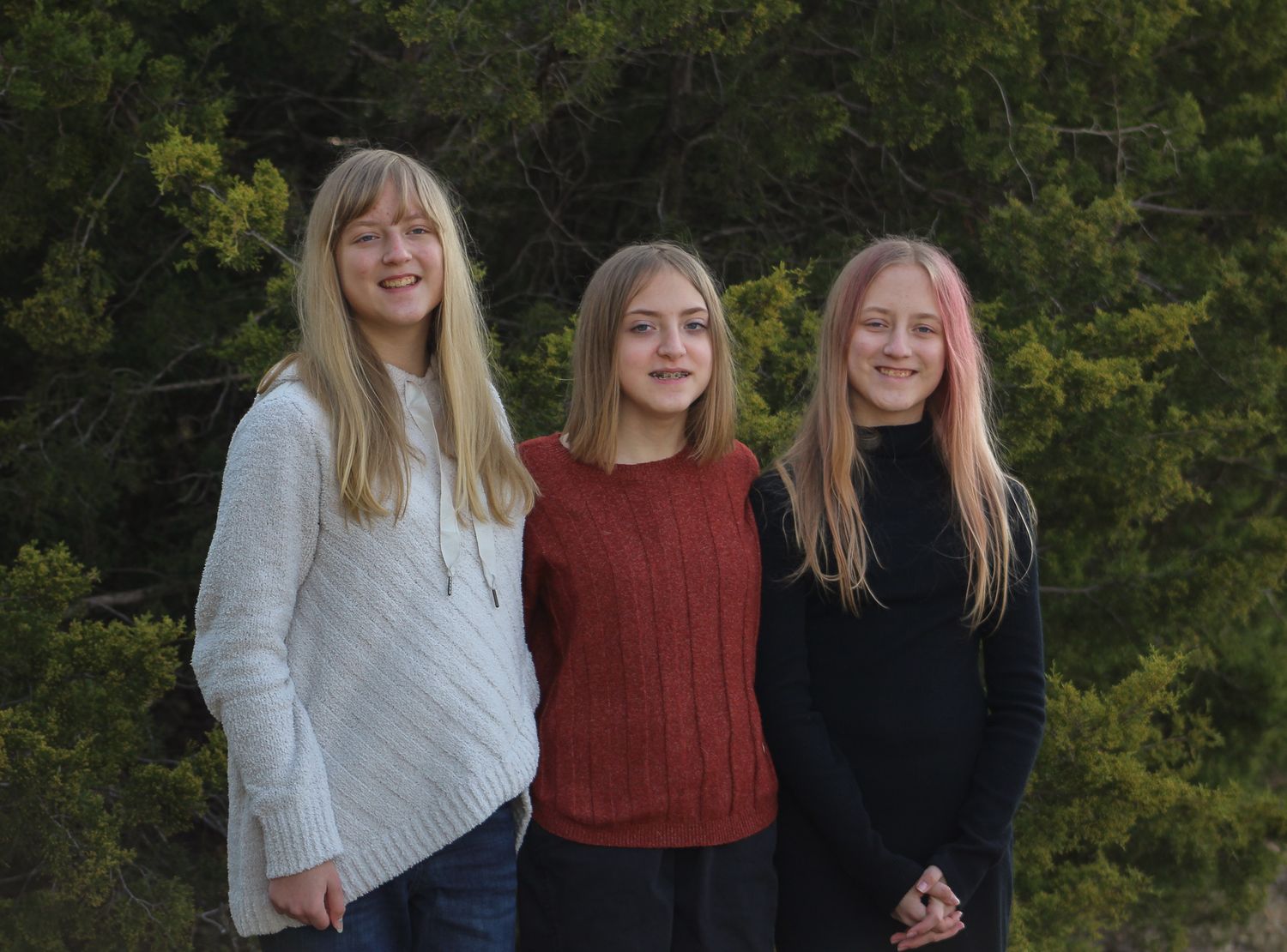 Three blonde girls smile together in an outdoor portrait with trees in the background.