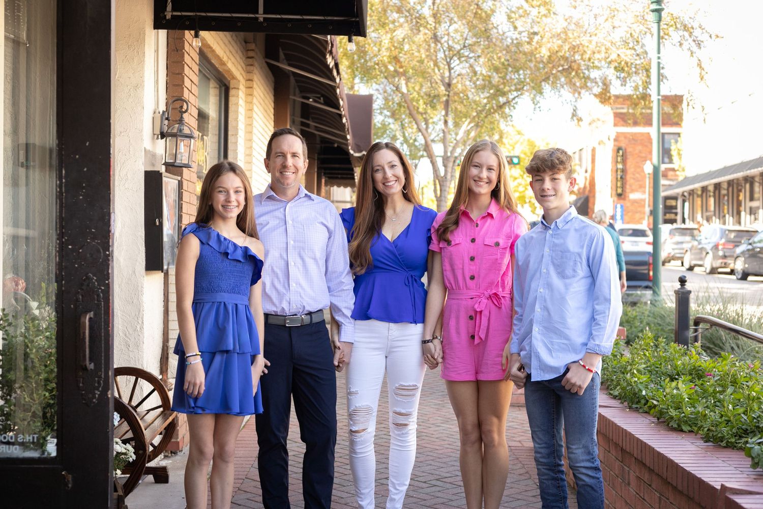 Family of five smiling together on a sunny sidewalk in coordinated blue and pink outfits.