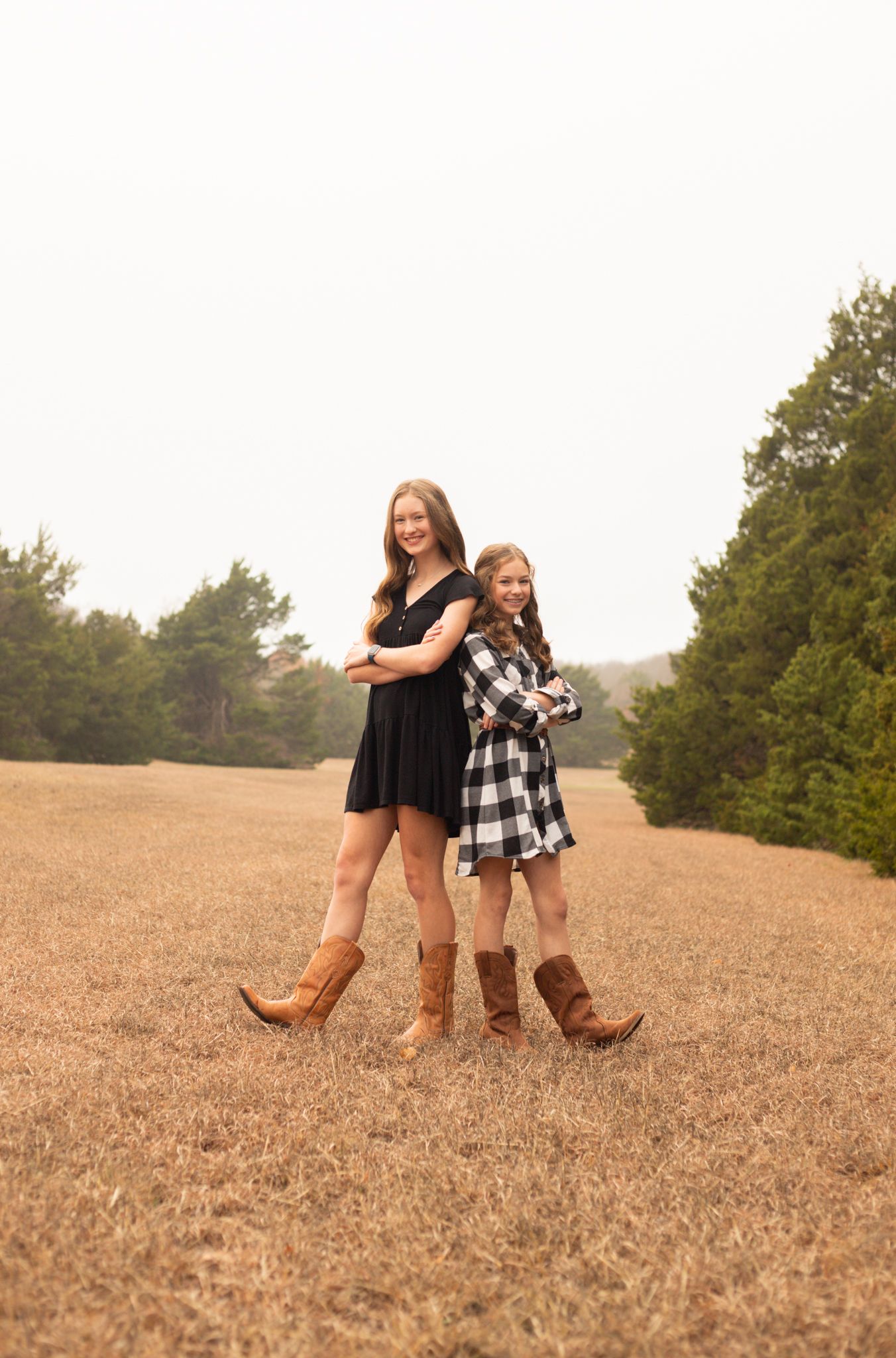 Two sisters posing back to back in a field wearing coordinating black and plaid outfits with cowboy boots.
