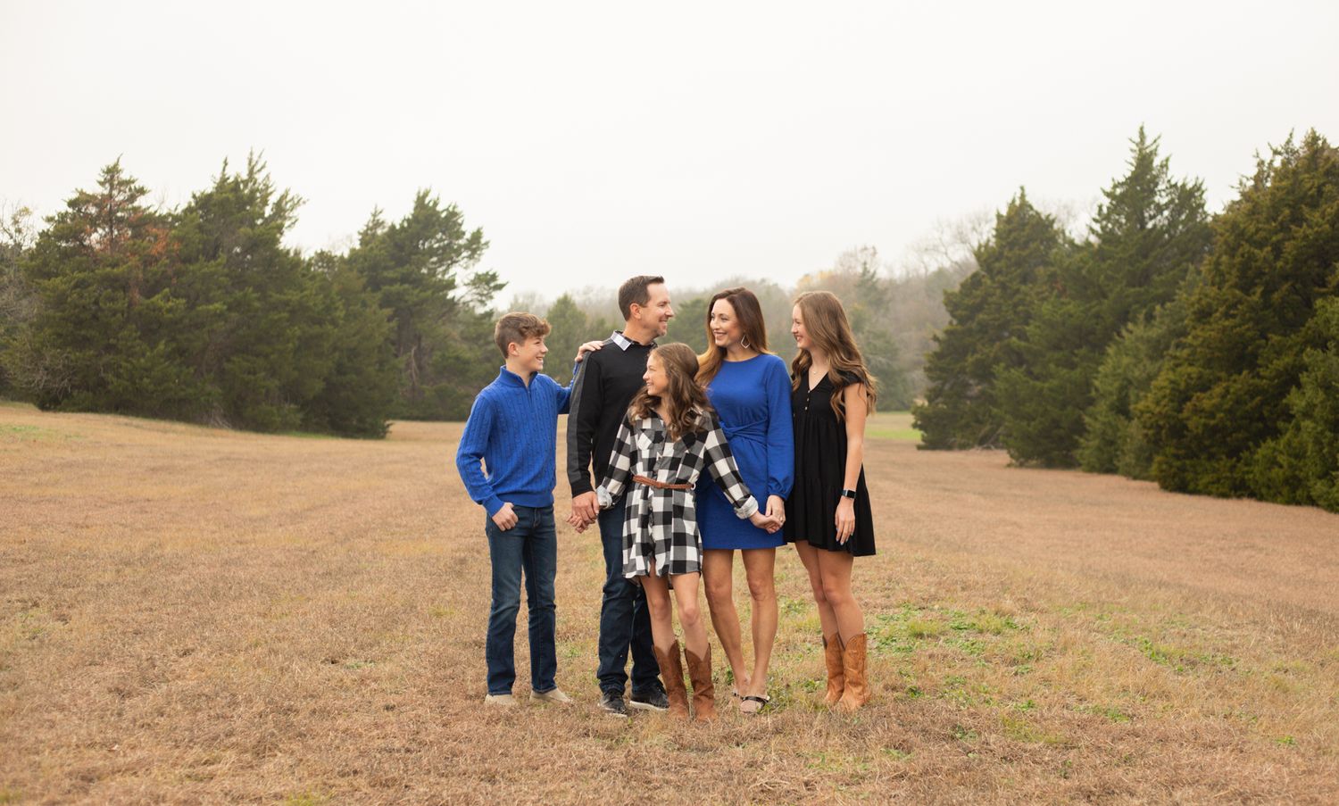 Family of five smiling together in an open field surrounded by trees during an outdoor photo session.