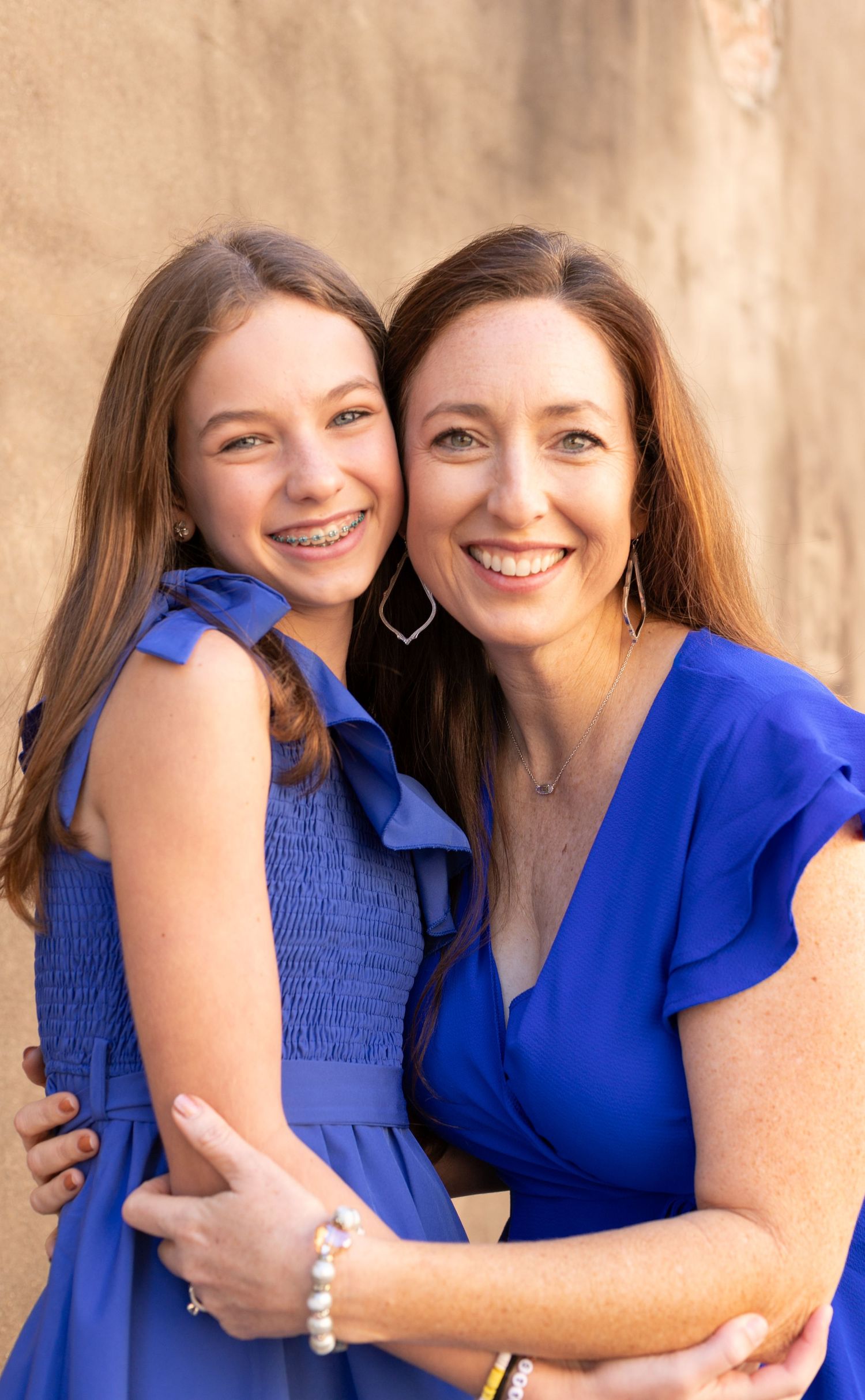 Smiling young girl with braces and woman in matching blue dresses embracing in a warm portrait.