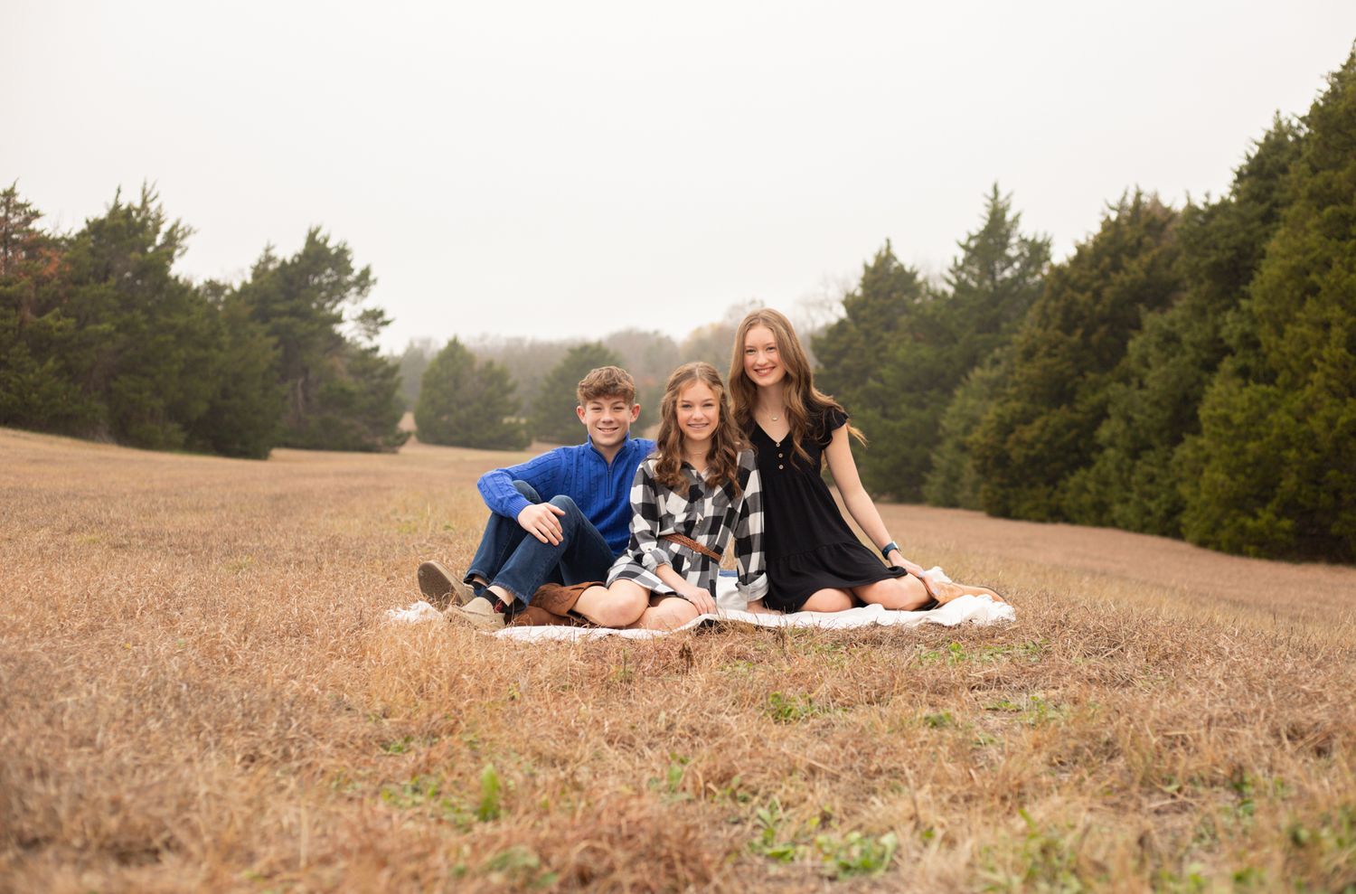 Three siblings sitting together on a blanket in a golden field for a family portrait session.