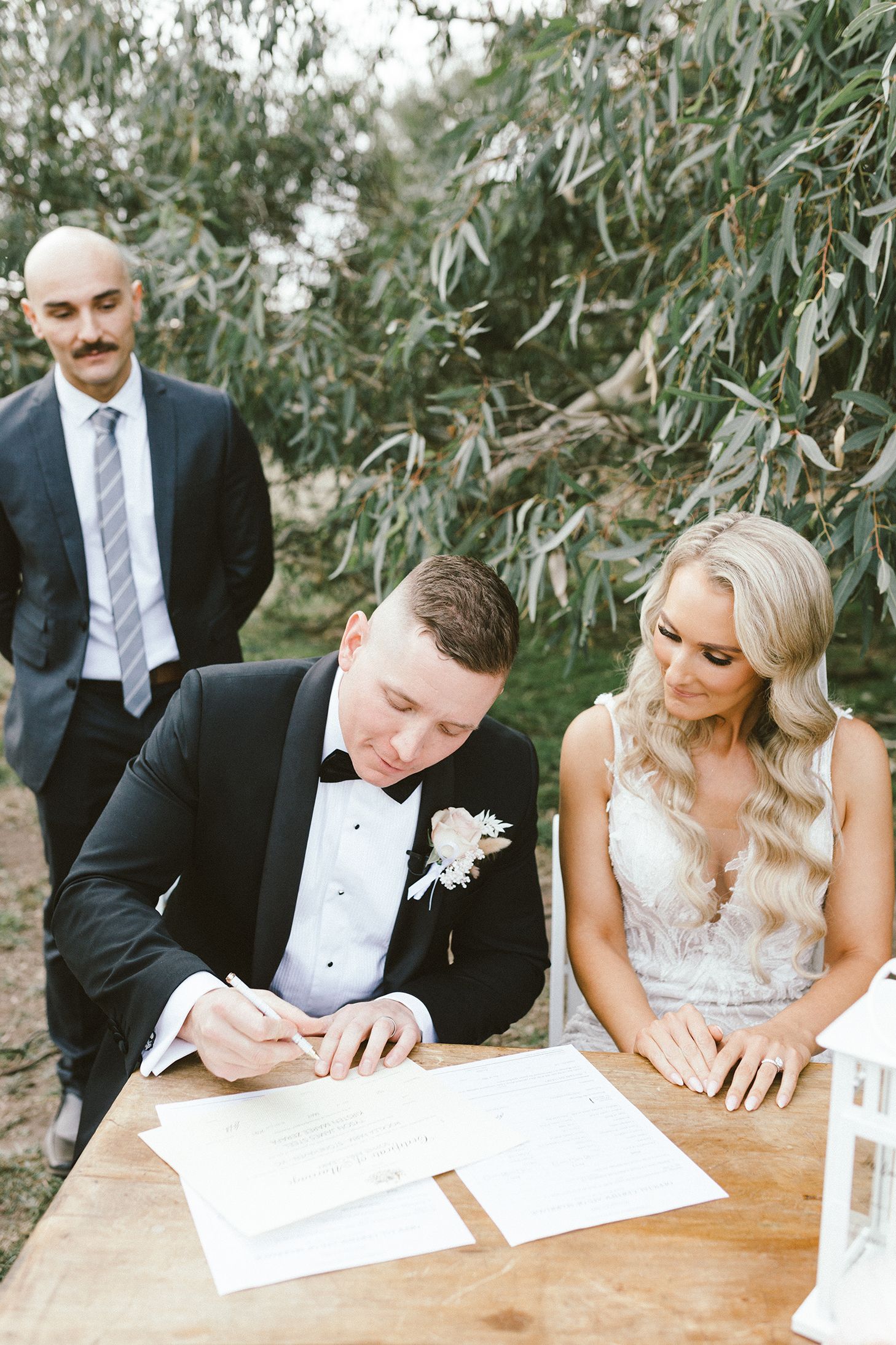 Wedding couple and witnesses signing official marriage documents at a rustic outdoor table.