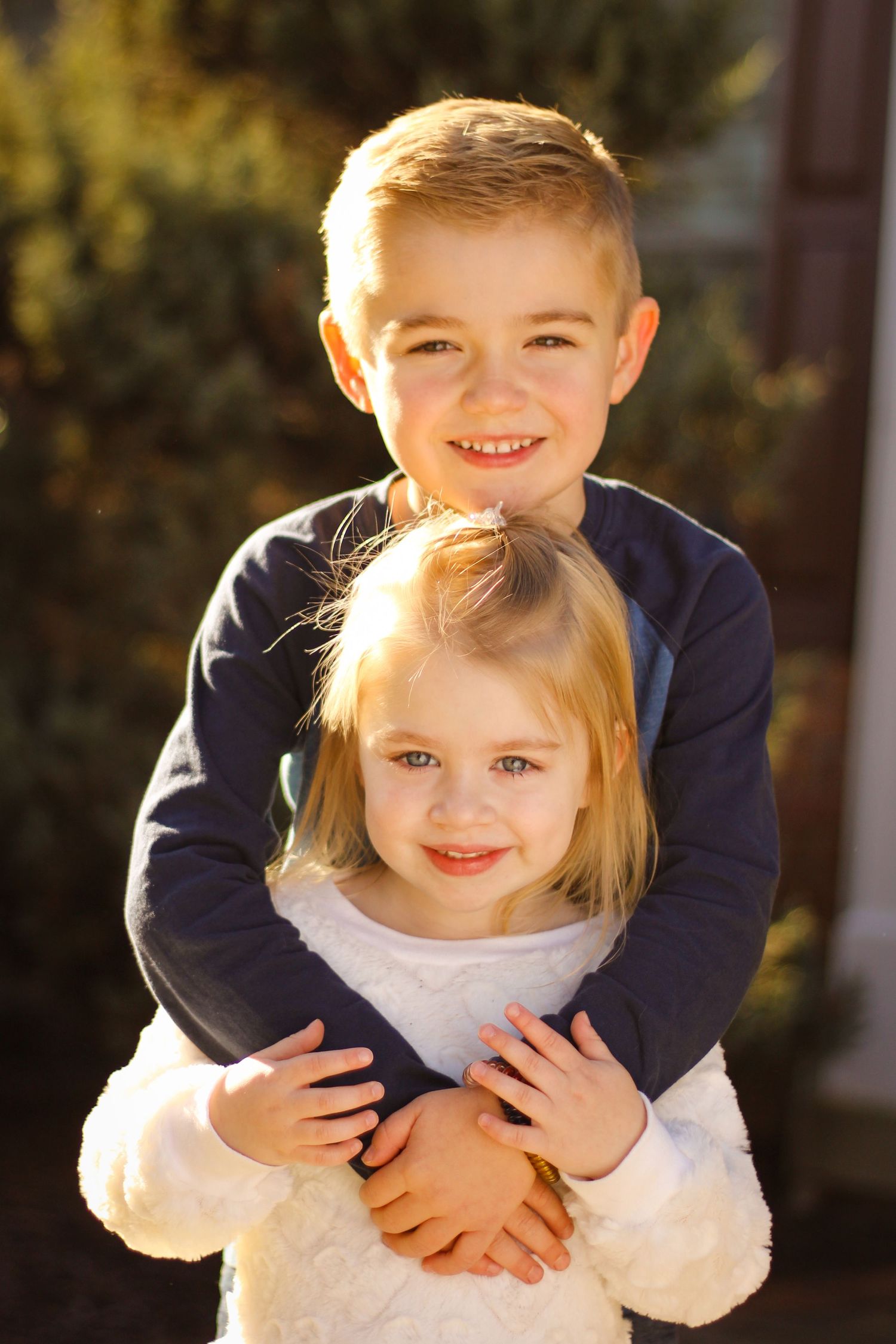 Two young children hugging and smiling outdoors in warm sunlight with one wearing navy and one wearing white.